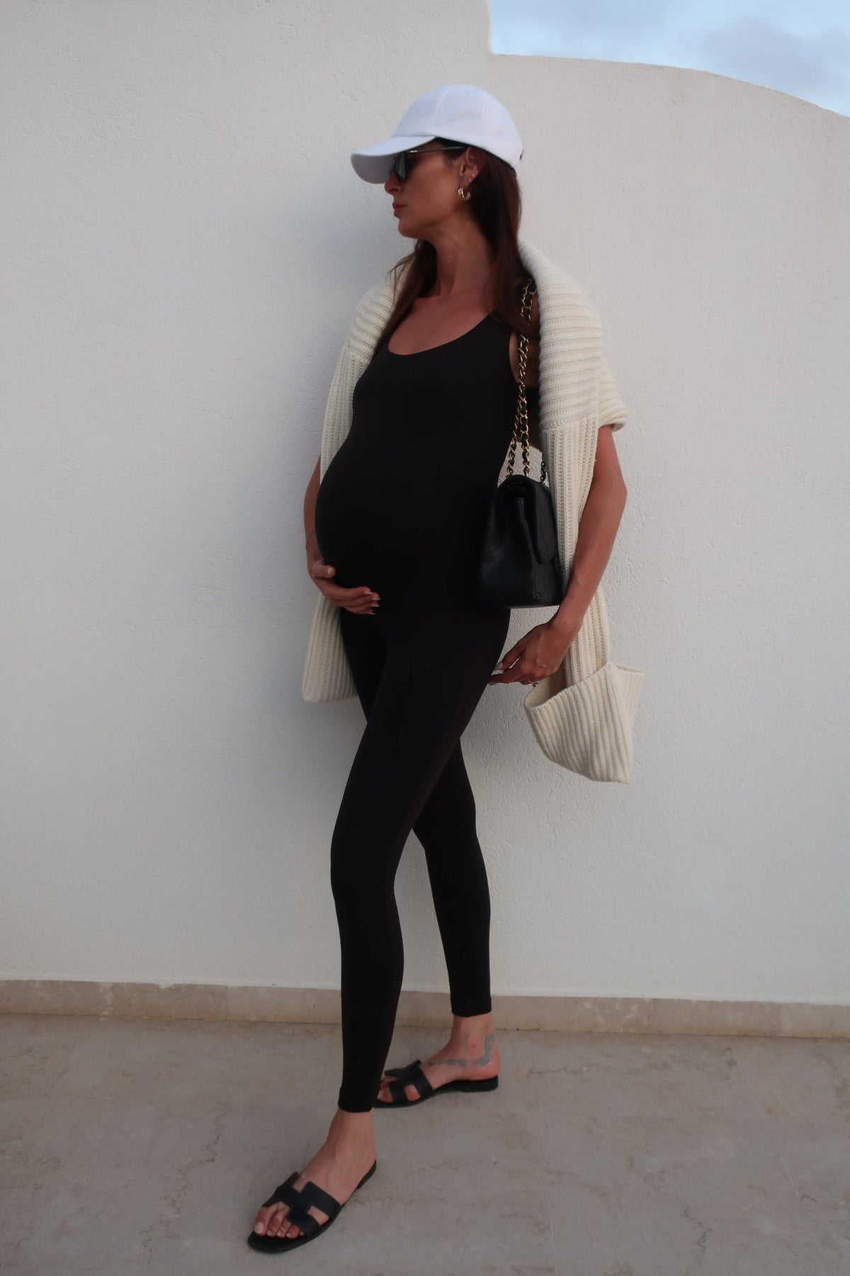 Full-length view of The Lucy Unitard in black showcasing its maternity fit on a pregnant model against a white wall