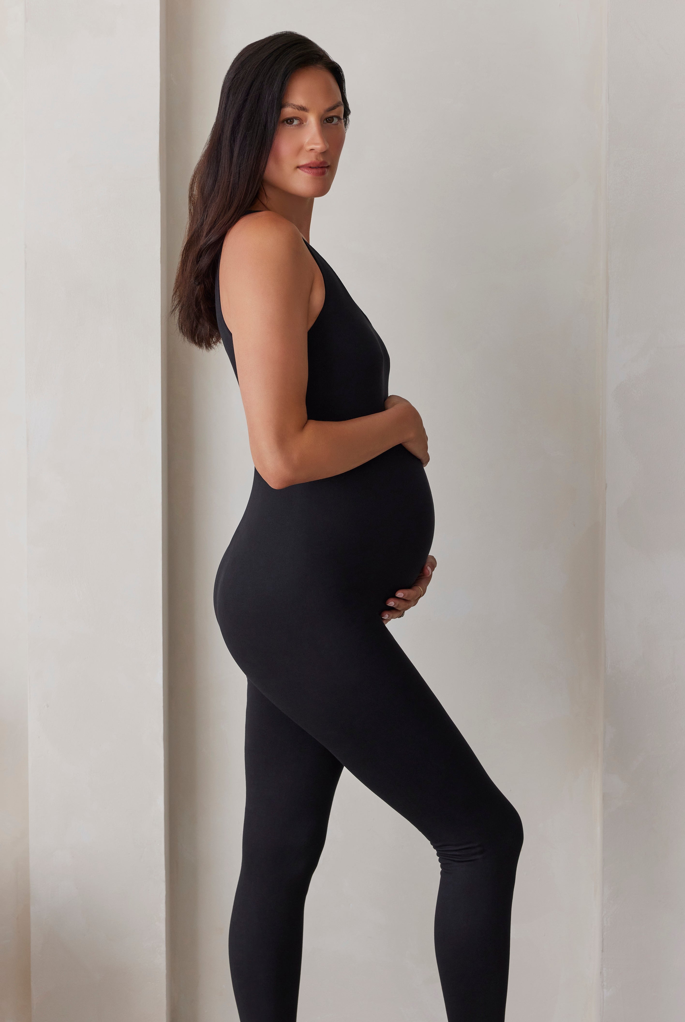 A pregnant woman wearing a black jumpsuit, standing in profile against a neutral beige wall.