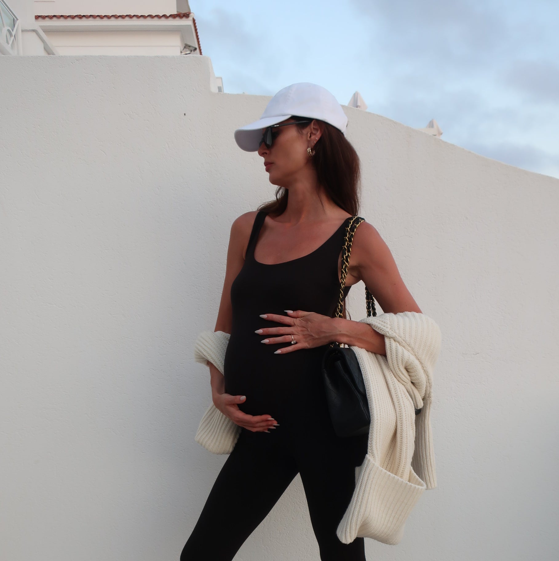 Pregnant woman in black bumpsuit and leggings holding a white jacket against a white wall.