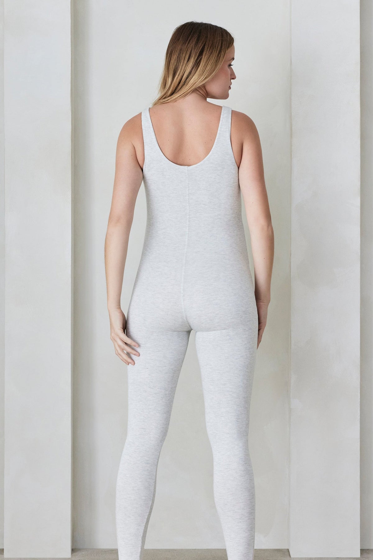 Back view of a woman wearing a heather grey Bumpsuit standing in front of a minimalist background with arches.