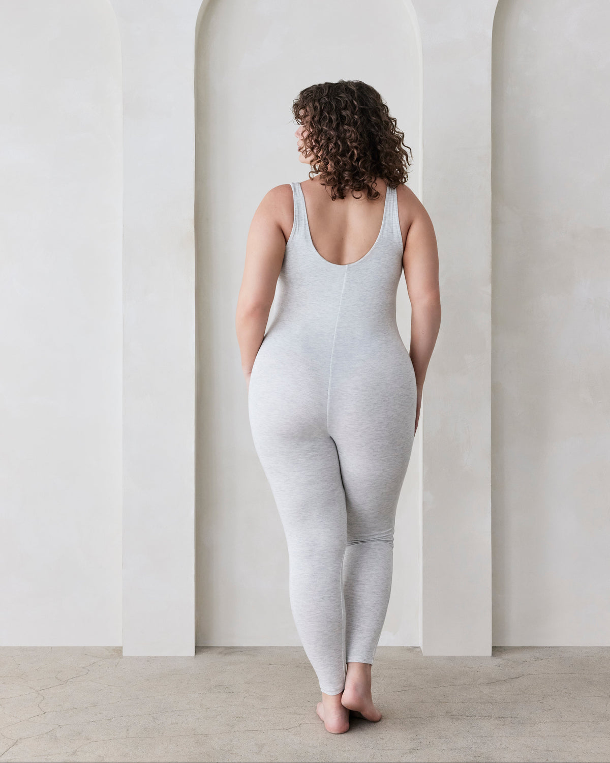 Woman wearing a grey bodysuit standing in a minimalistic room with white walls and arches.