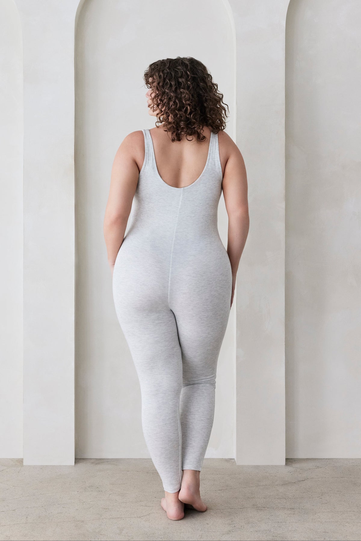Woman wearing a grey bodysuit standing in a minimalistic room with white walls and arches.