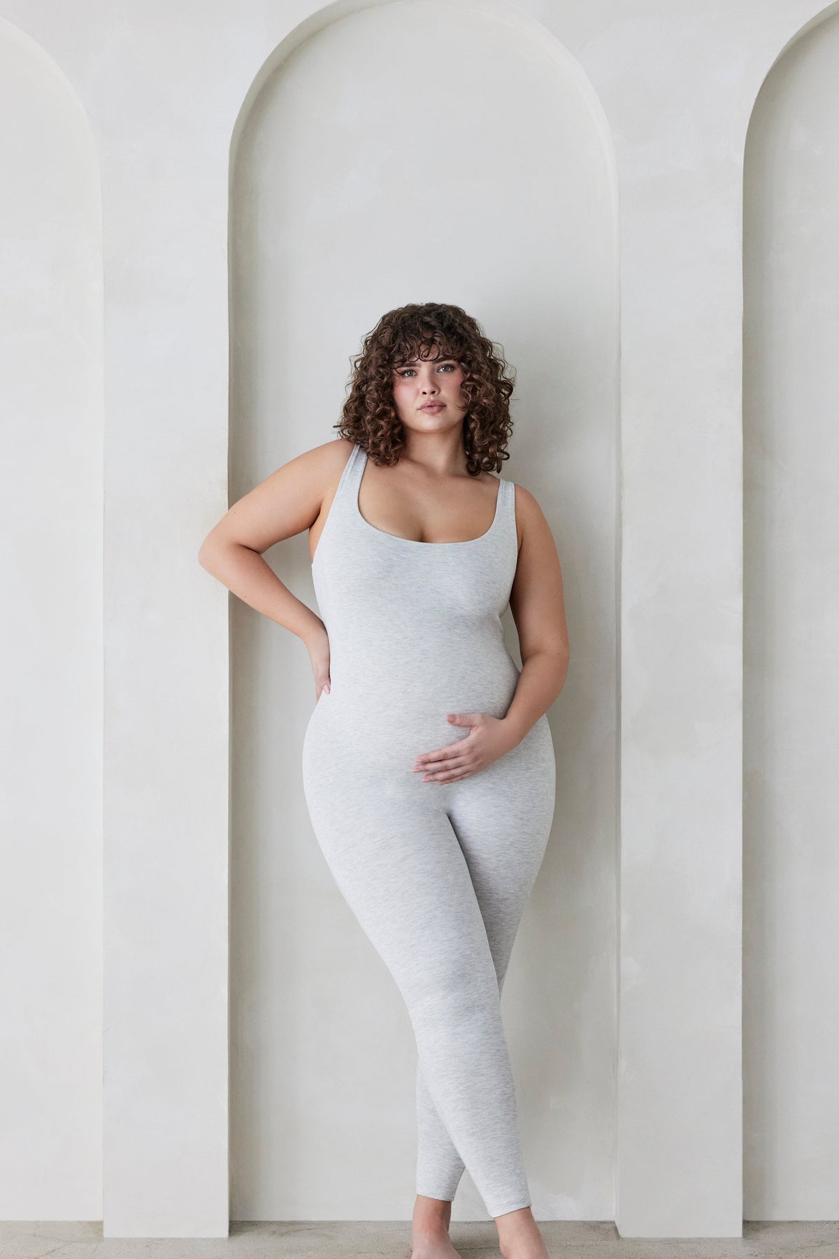 Pregnant woman wearing the Lucy maternity scoop neck unitard in heather grey by Bumpsuit set against a feature wall