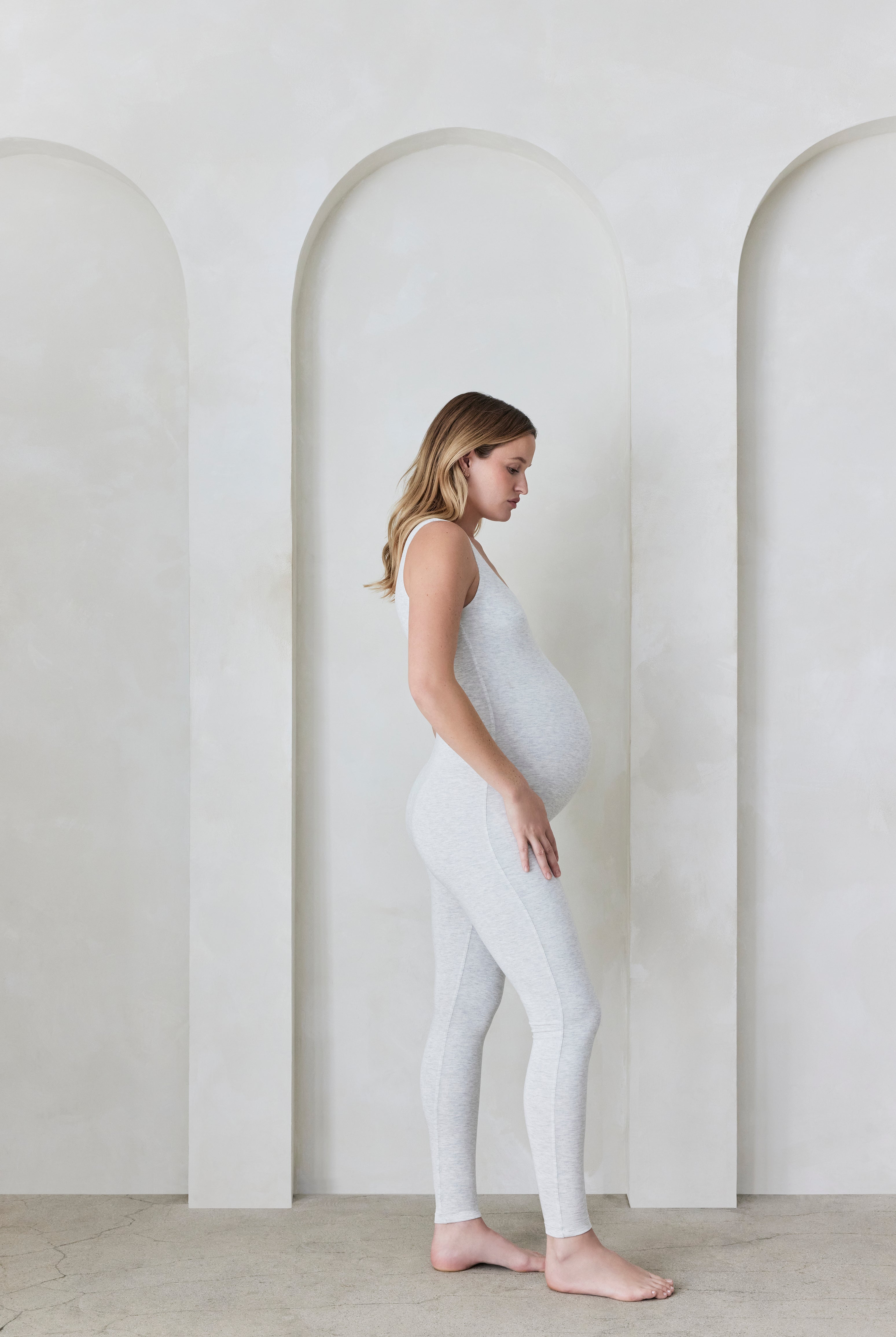 Pregnant woman wearing a heather grey Bumpsuit in a minimalistic setting with arches.