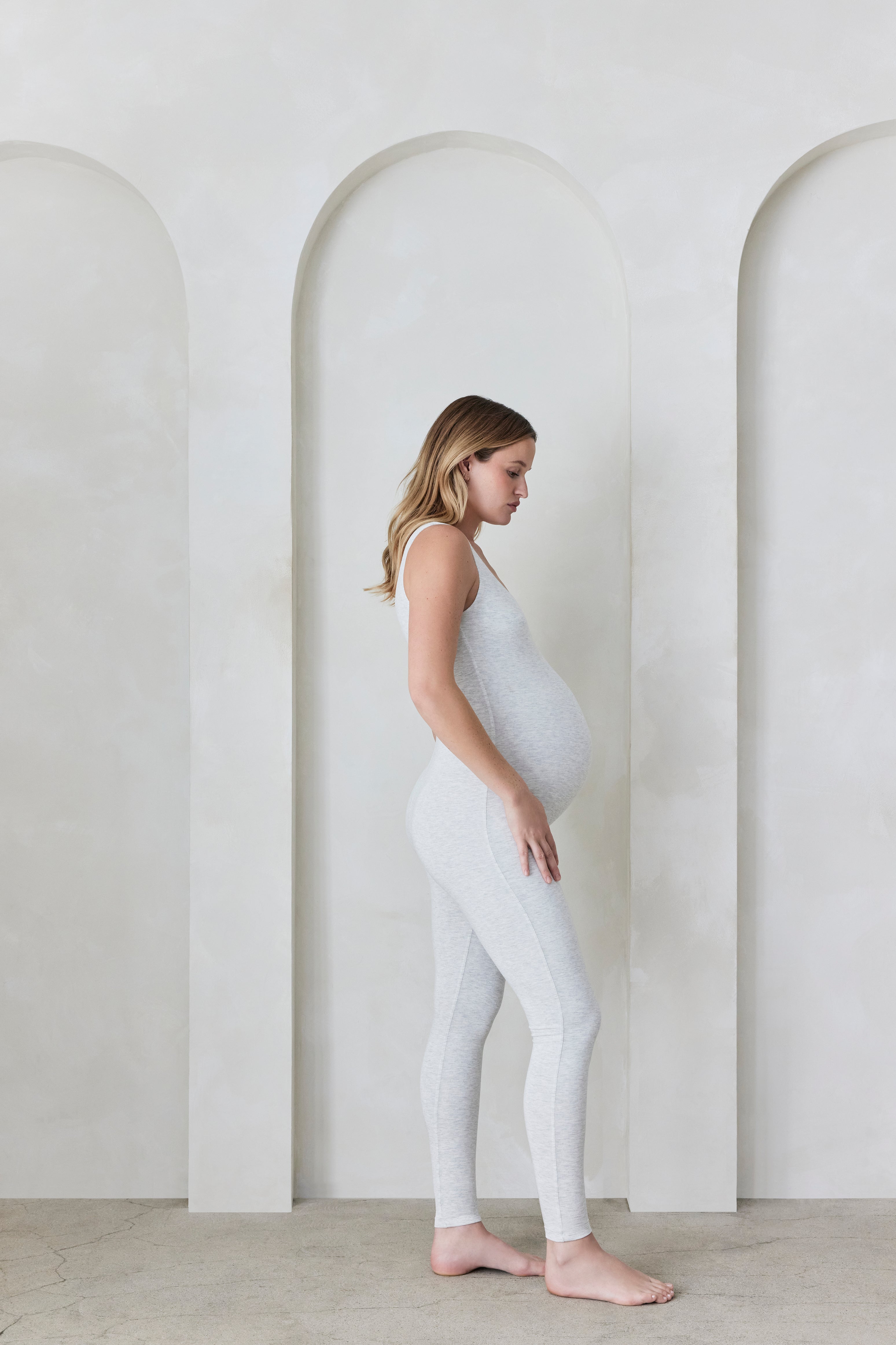 Pregnant woman wearing a heather grey Bumpsuit in a minimalistic setting with arches.