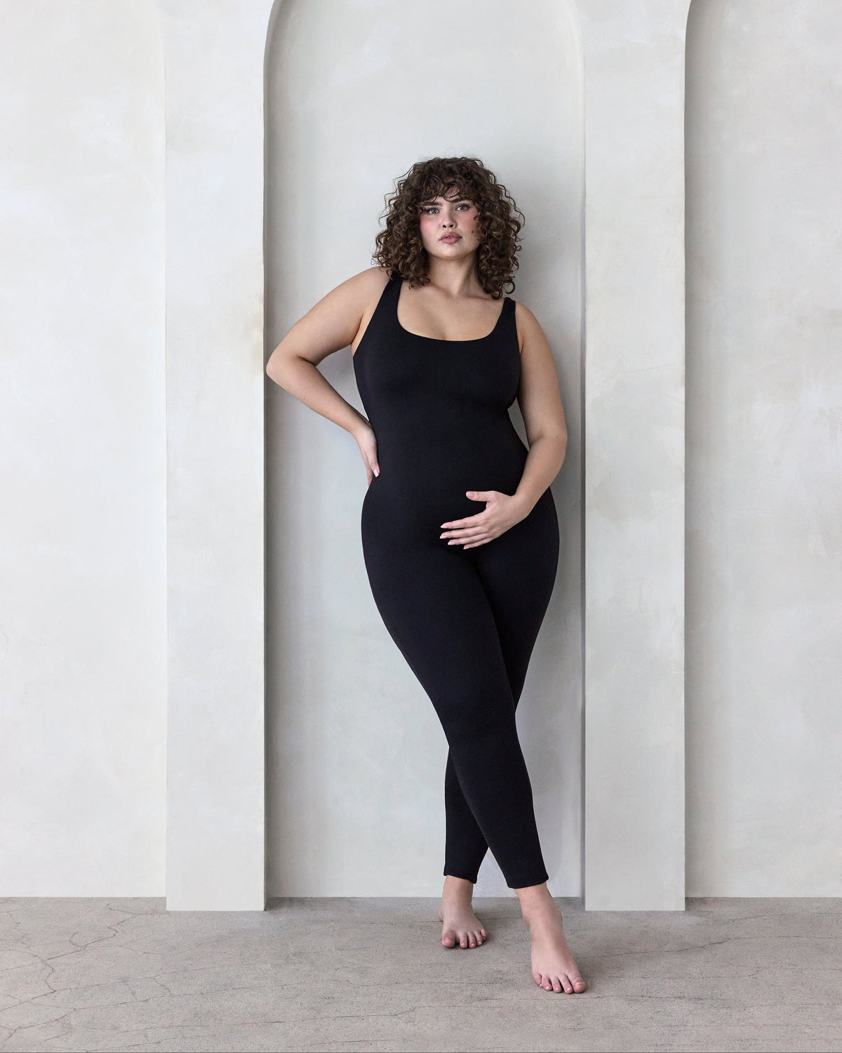 Woman wearing a black Bumpsuit Jumpsuit standing in a minimalistic setting with arches.