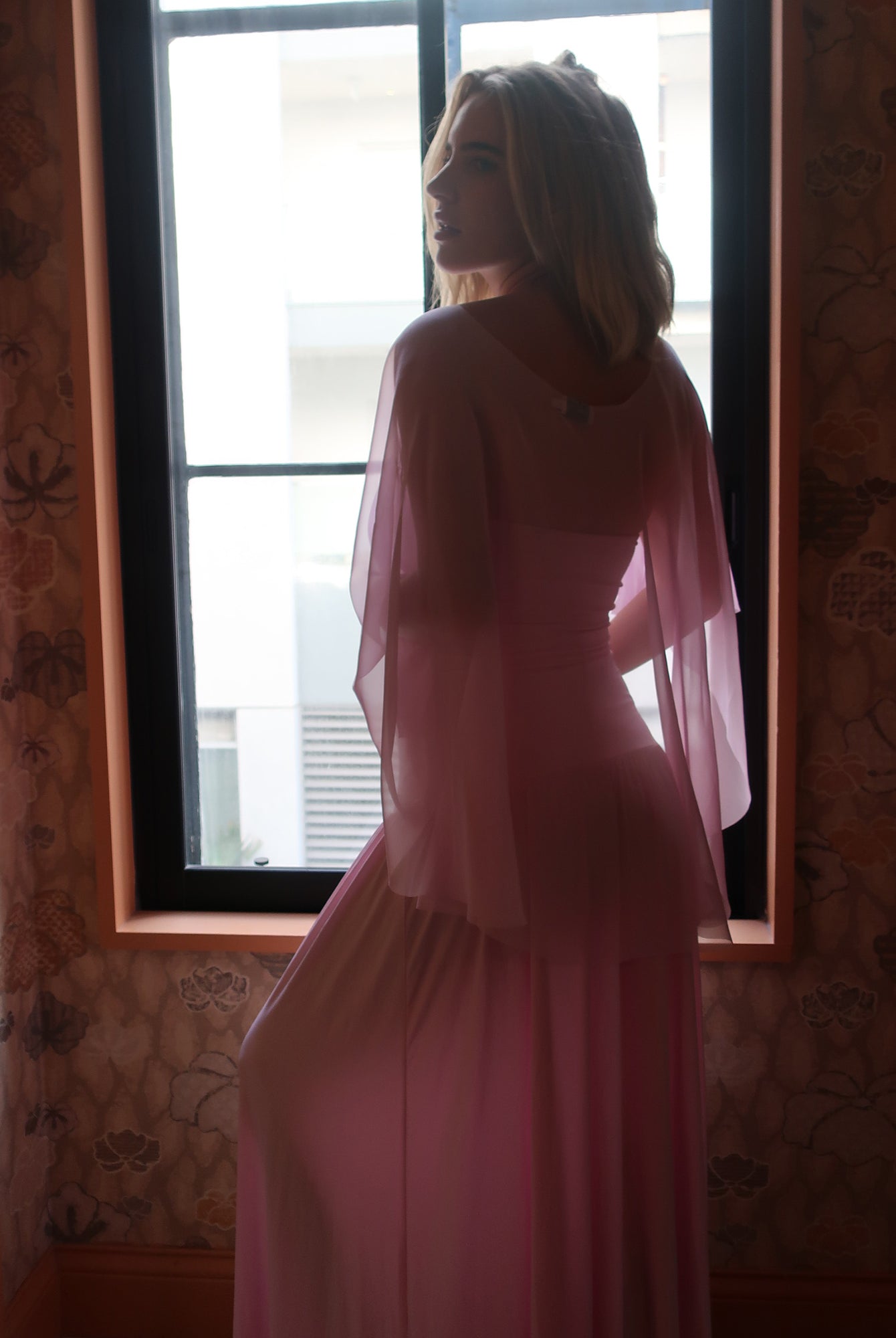 A pregnant woman stands in profile by a window wearing a flowing pink dress and chiffon cape by Bumpsuit , photographed in a dimly lit interior with patterned wallpaper.