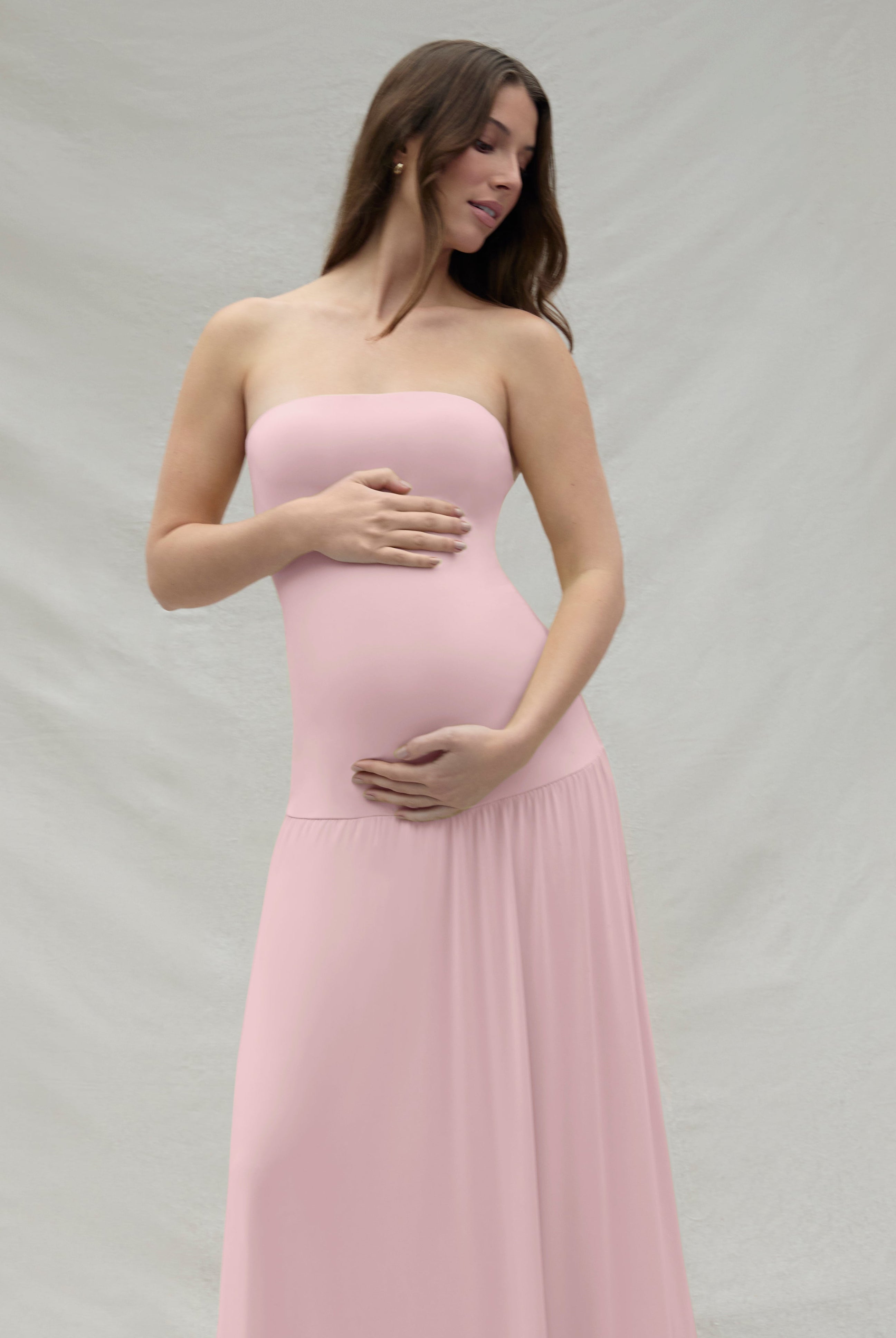 Pregnant woman wearing a pink strapless gown by Bumpsuit against a beige background