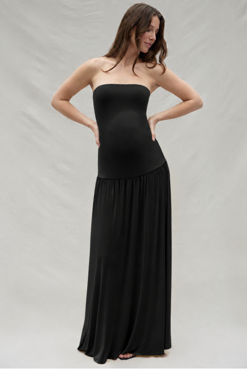 Pregnant woman wearing a Bumpsuit strapless maxi dress with a drop waist feature, standing in-front of an off white backdrop