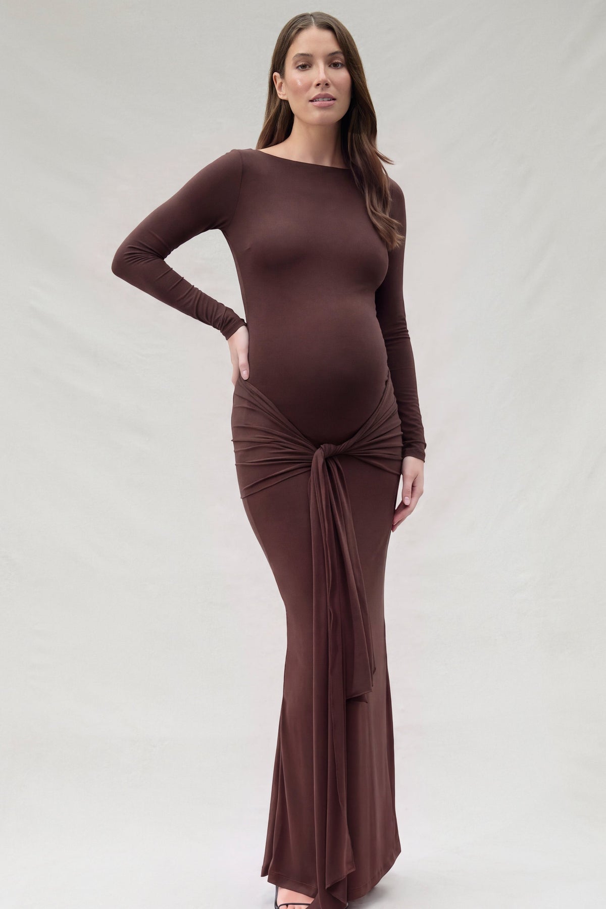 A pregnant woman modeling a long-sleeve, chocolate brown maternity maxi dress with a gathered tie detail at the waist, set against a plain beige studio background.