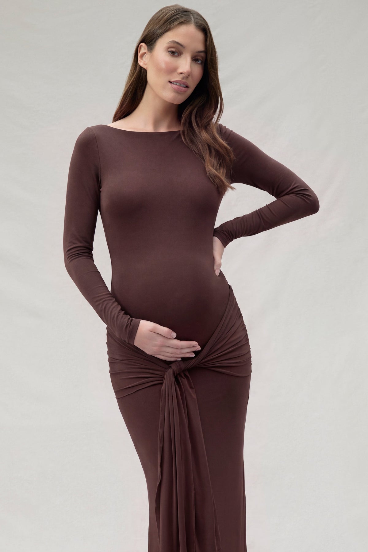 A pregnant woman modeling a long-sleeve, chocolate brown Bumpsuit maternity dress with a gathered twist tie detail at the waist, set against a neutral beige studio backdrop.