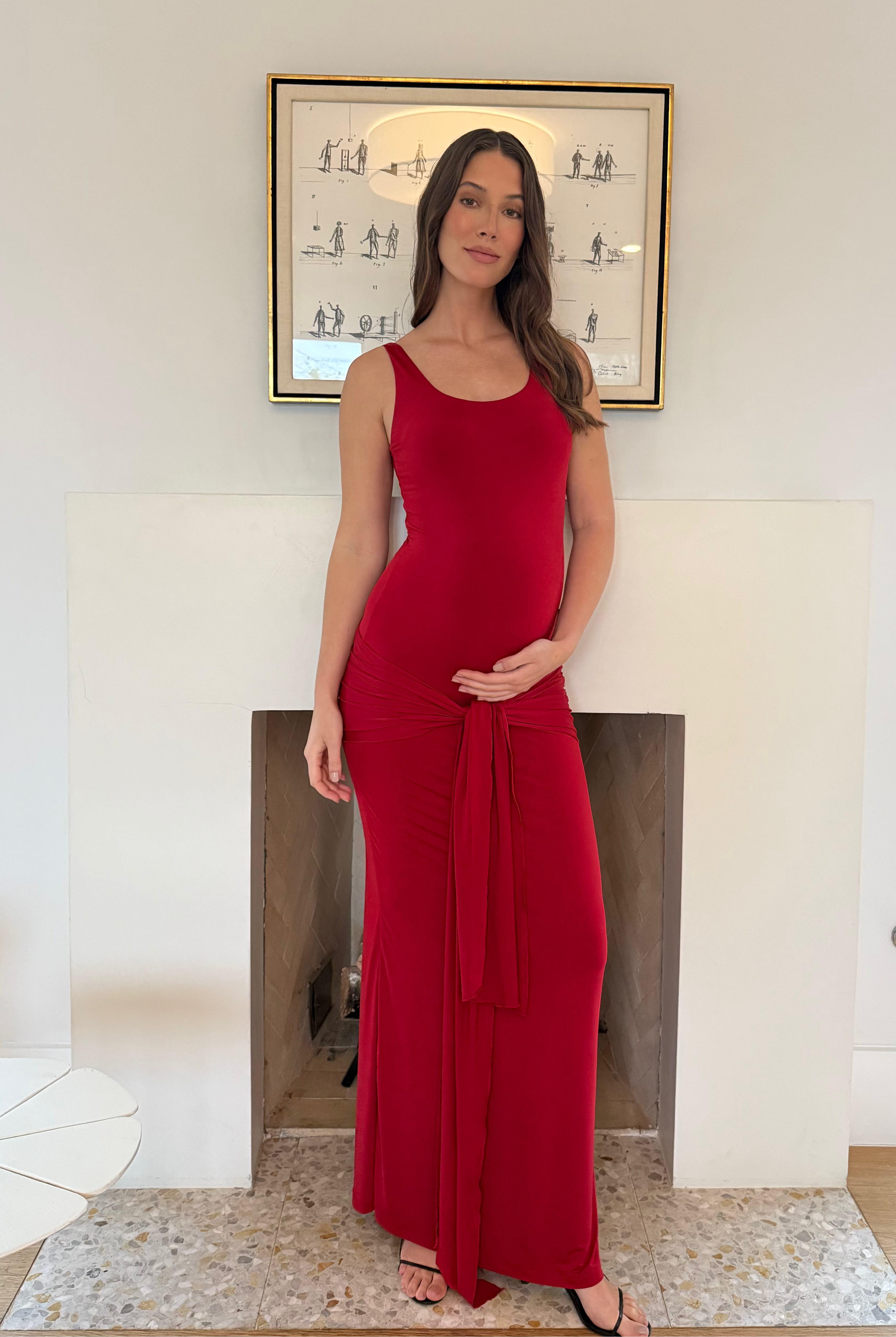Pregnant woman in a Bumpsuit red maternity dress standing in a room with a fireplace and framed picture on the wall.