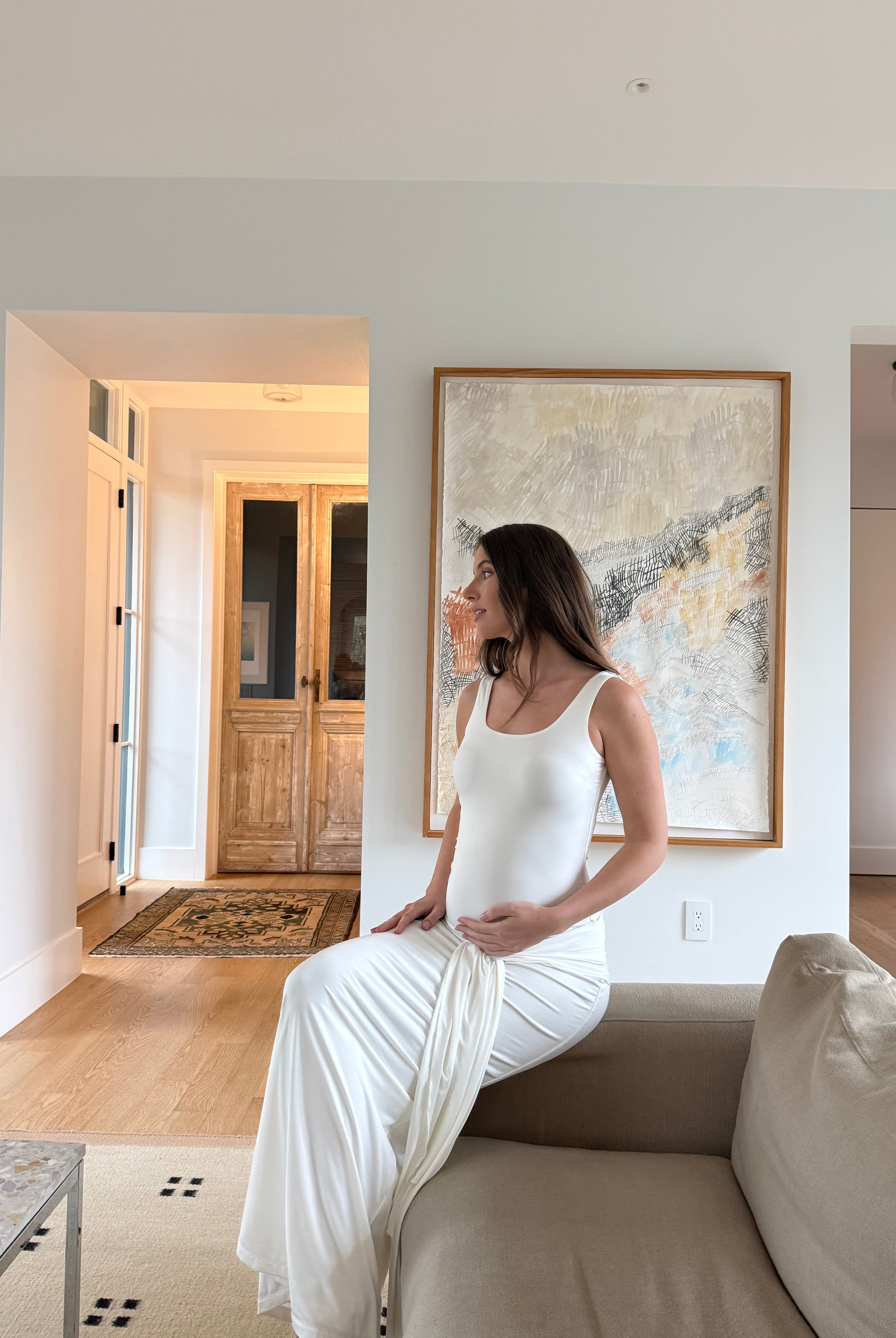 Pregnant woman in a white dress sitting on a couch in a modern living room with abstract art on the wall.