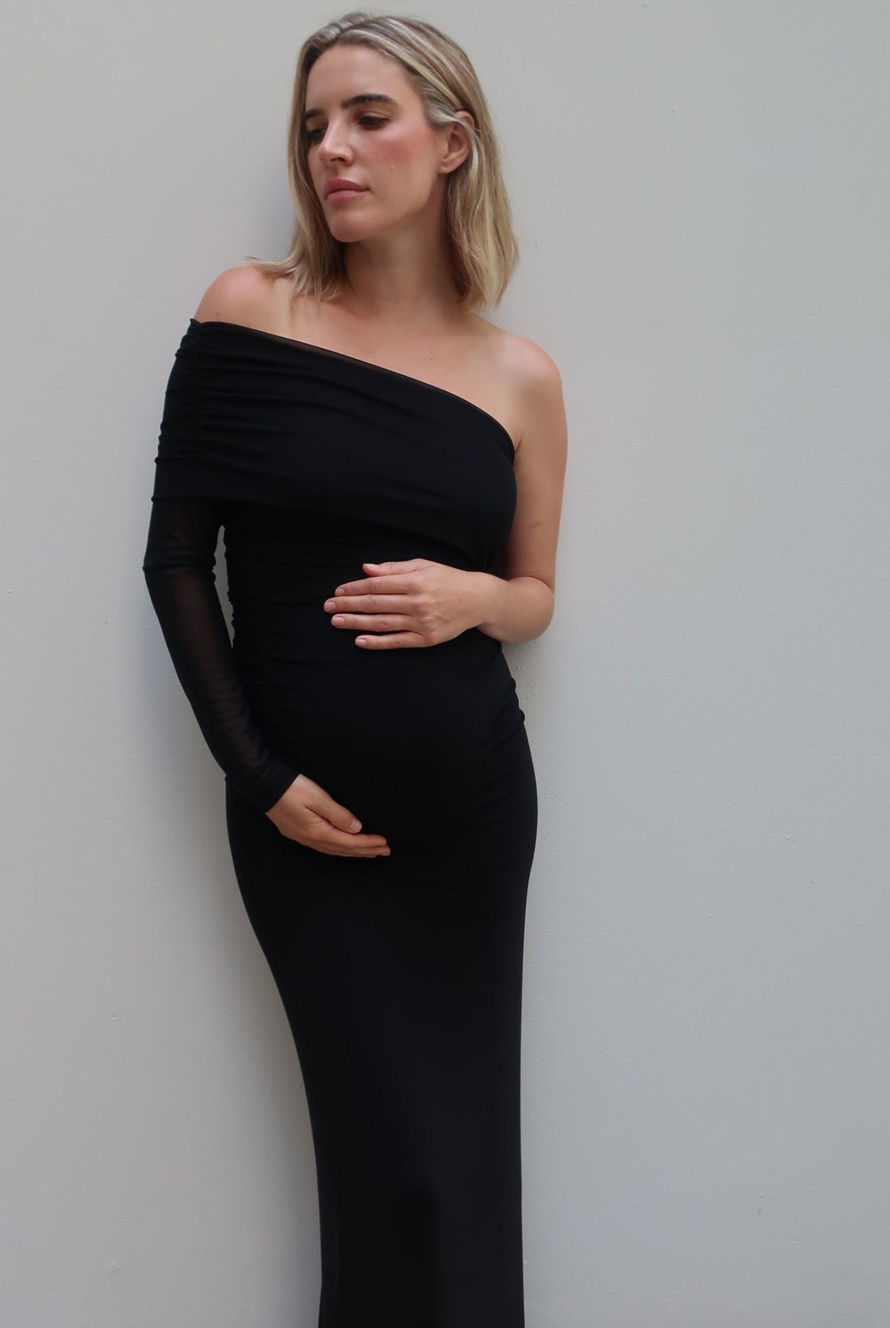 A woman wearing a Bumpsuit black off-the-shoulder long-sleeve maternity gown, standing against a plain light gray wall.