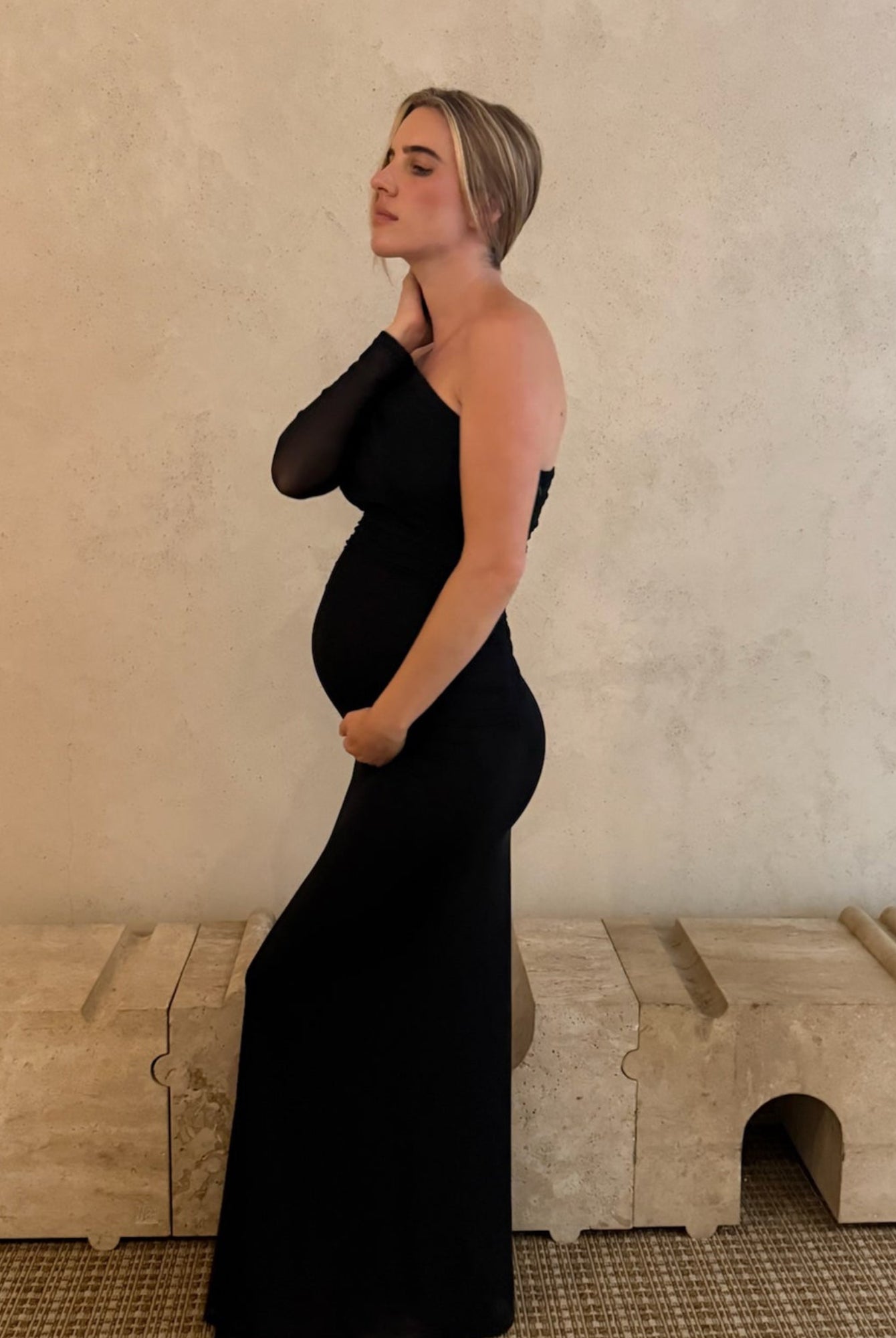 A pregnant woman wearing a black one-shoulder maternity gown by Bumpsuit stands in profile against a textured beige wall, with a light stone bench and woven rug in the background.