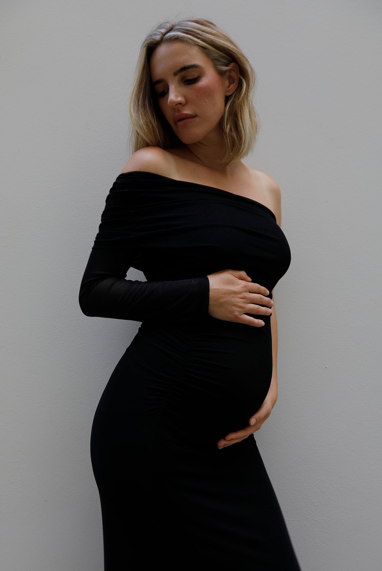 A pregnant woman wearing a Bumpsuit black off-the-shoulder long-sleeve maternity dress, posing with her hands on her belly against a plain gray wall.