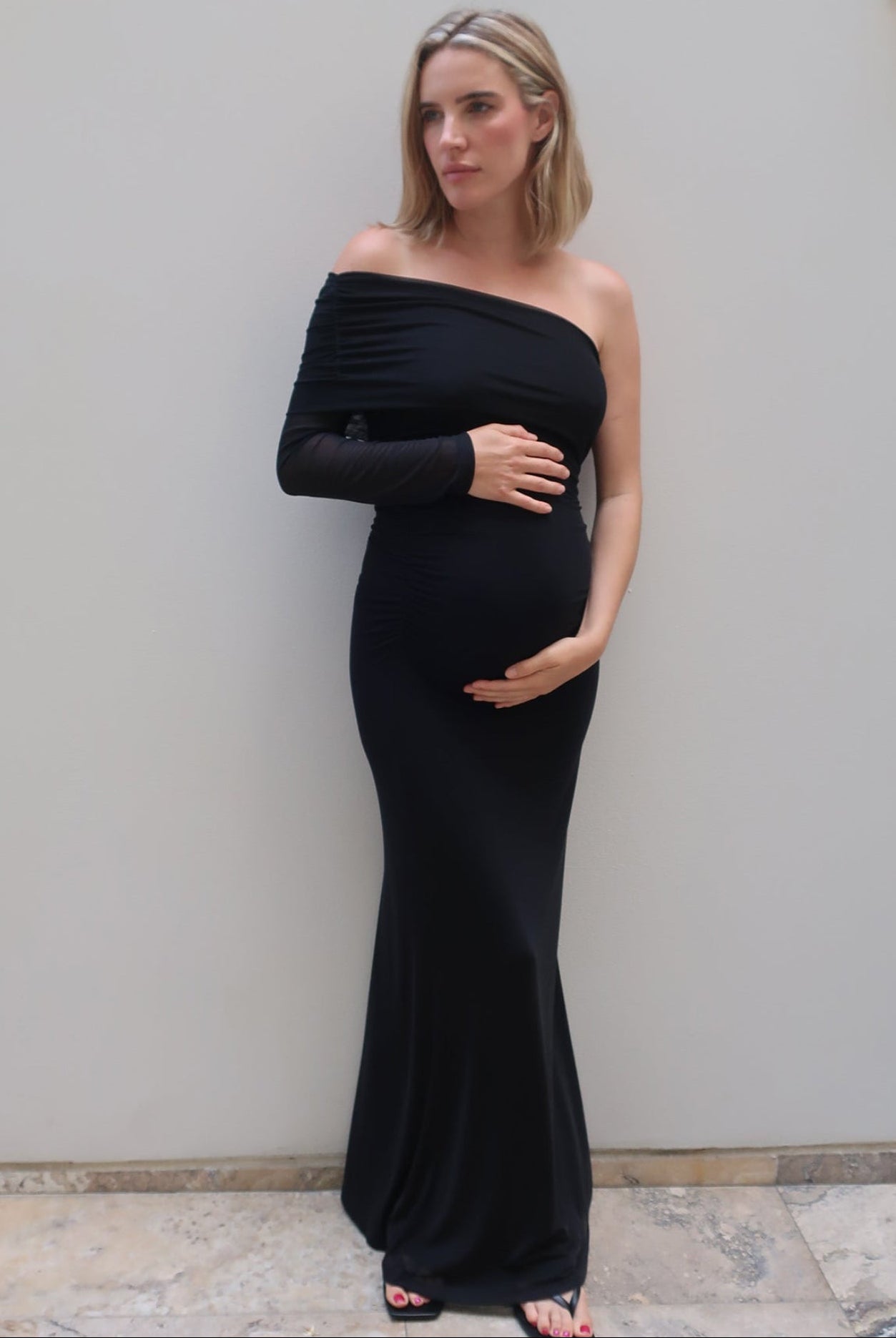A pregnant woman wearing a black off-the-shoulder long-sleeve maternity gown by Bumpsuit, standing against a light gray wall.