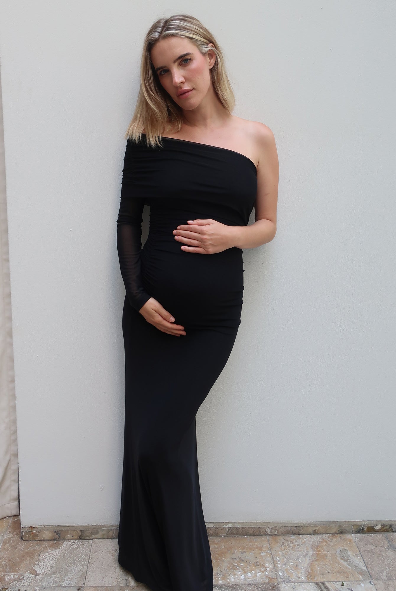 A pregnant woman with blonde hair models a black one-shoulder maternity gown with sheer long sleeves by Bumpsuit, posing against a plain light gray wall.