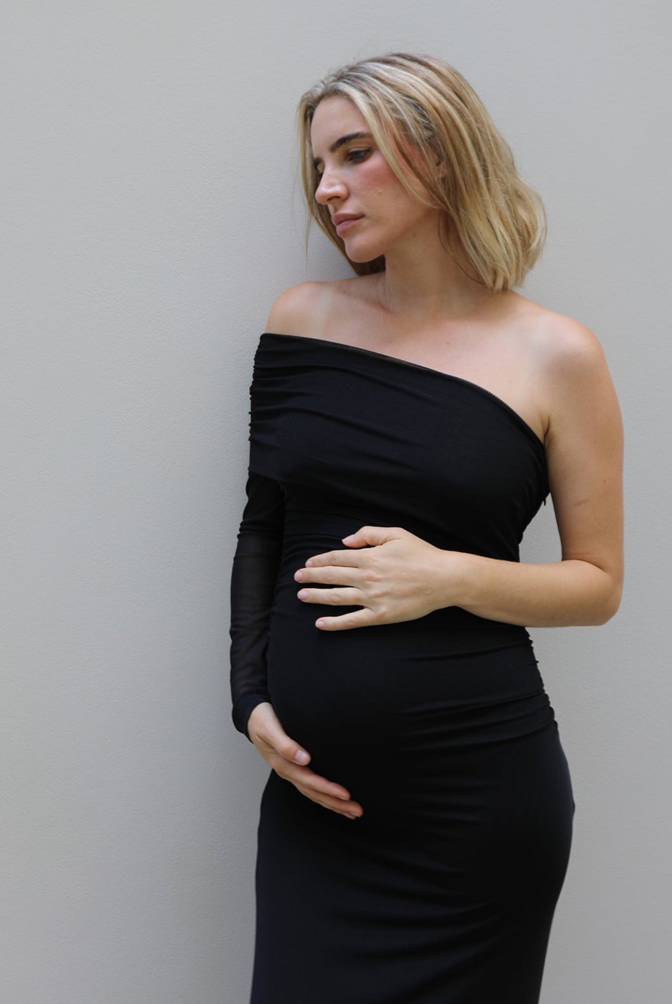 A pregnant woman with blonde hair wearing a black off-the-shoulder long-sleeve maternity gown by Bumpsuit, posing with her hands on her belly against a light gray wall.