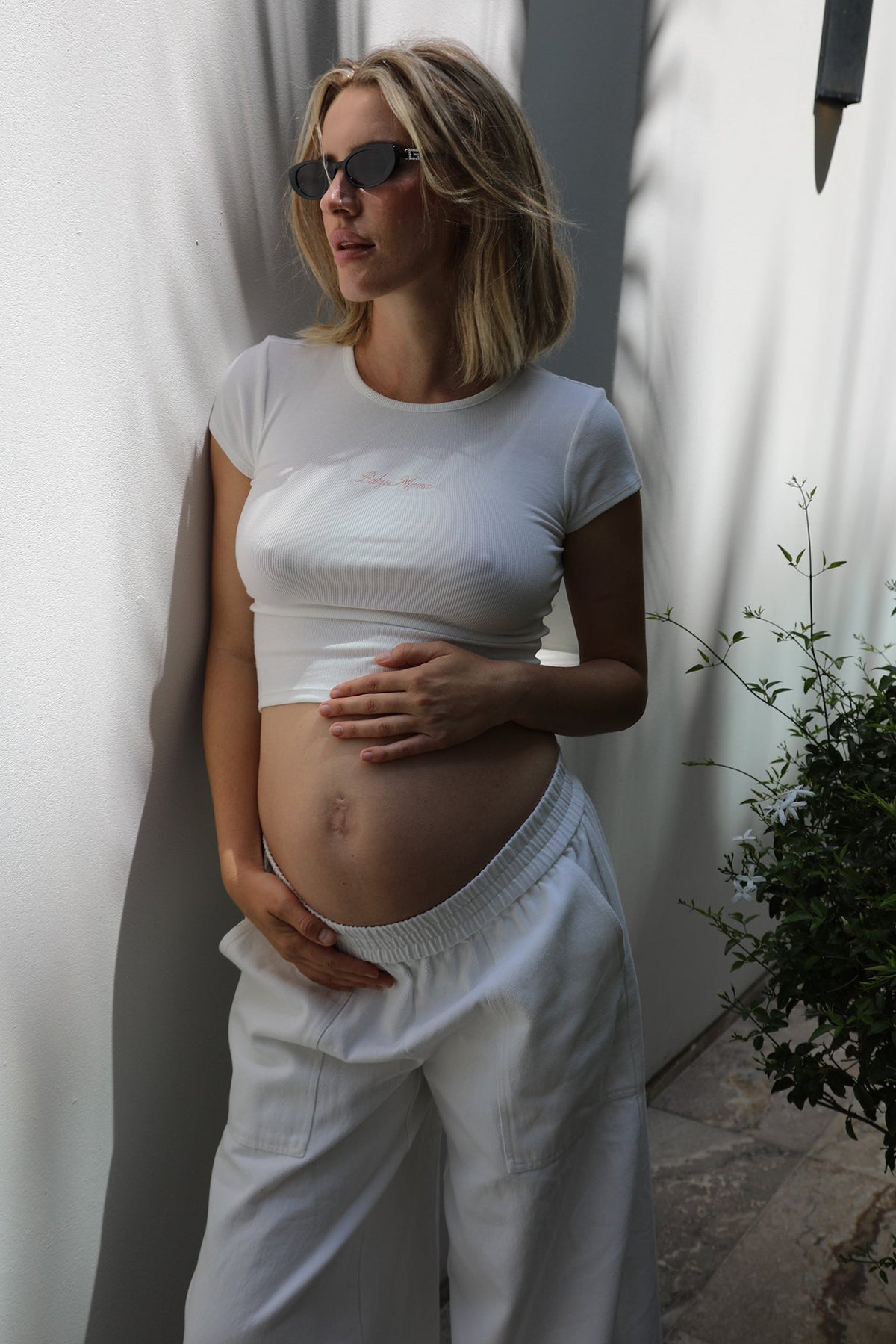A pregnant woman wearing a white short-sleeve crop top and white wide leg pants, standing in a sunlit outdoor setting with shadows on a white wall.