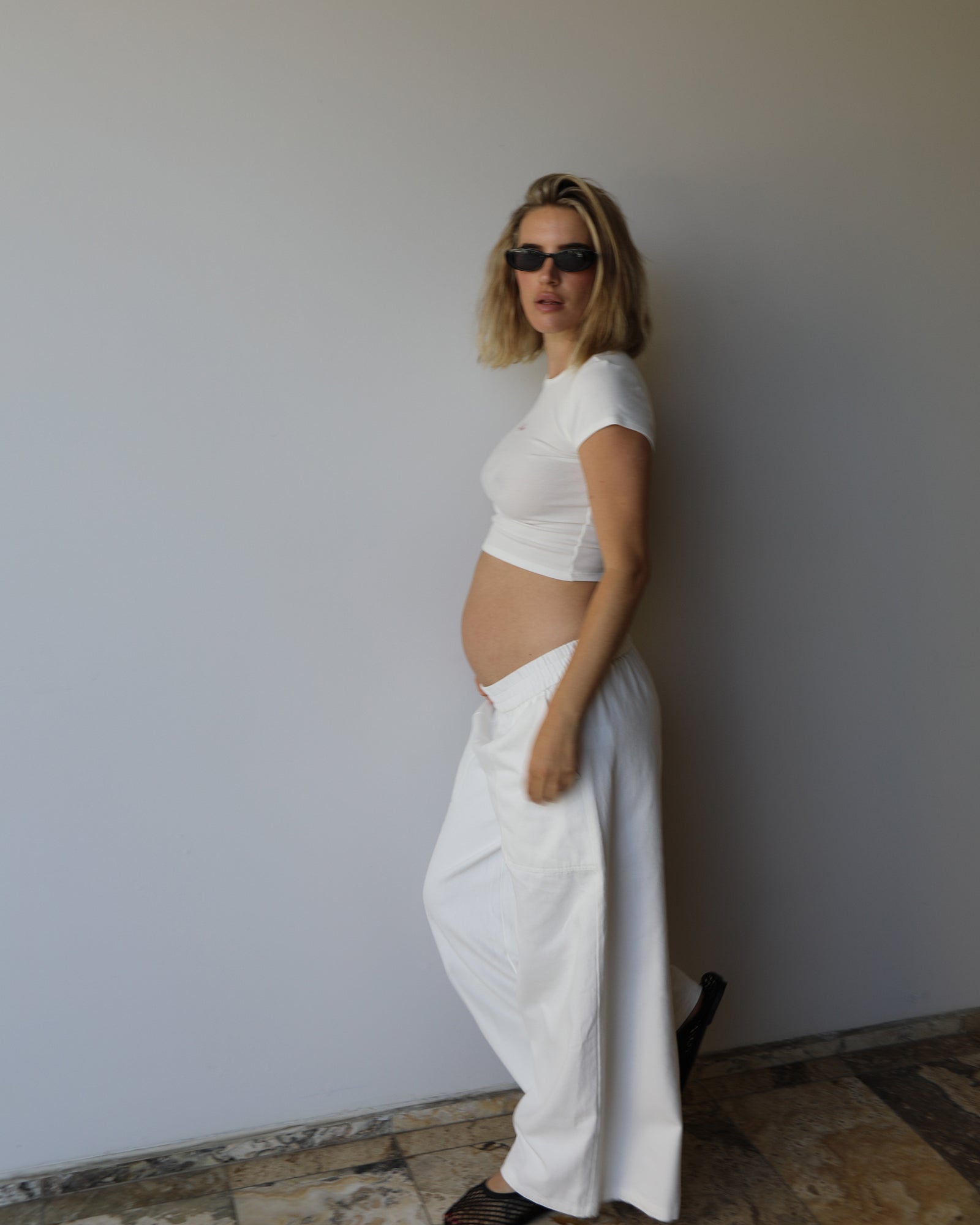 A pregnant woman with blonde hair and sunglasses stands in profile wearing a ivory short-sleeve crop top and matching white wide-leg pants by Bumpsuit against a plain light gray wall.