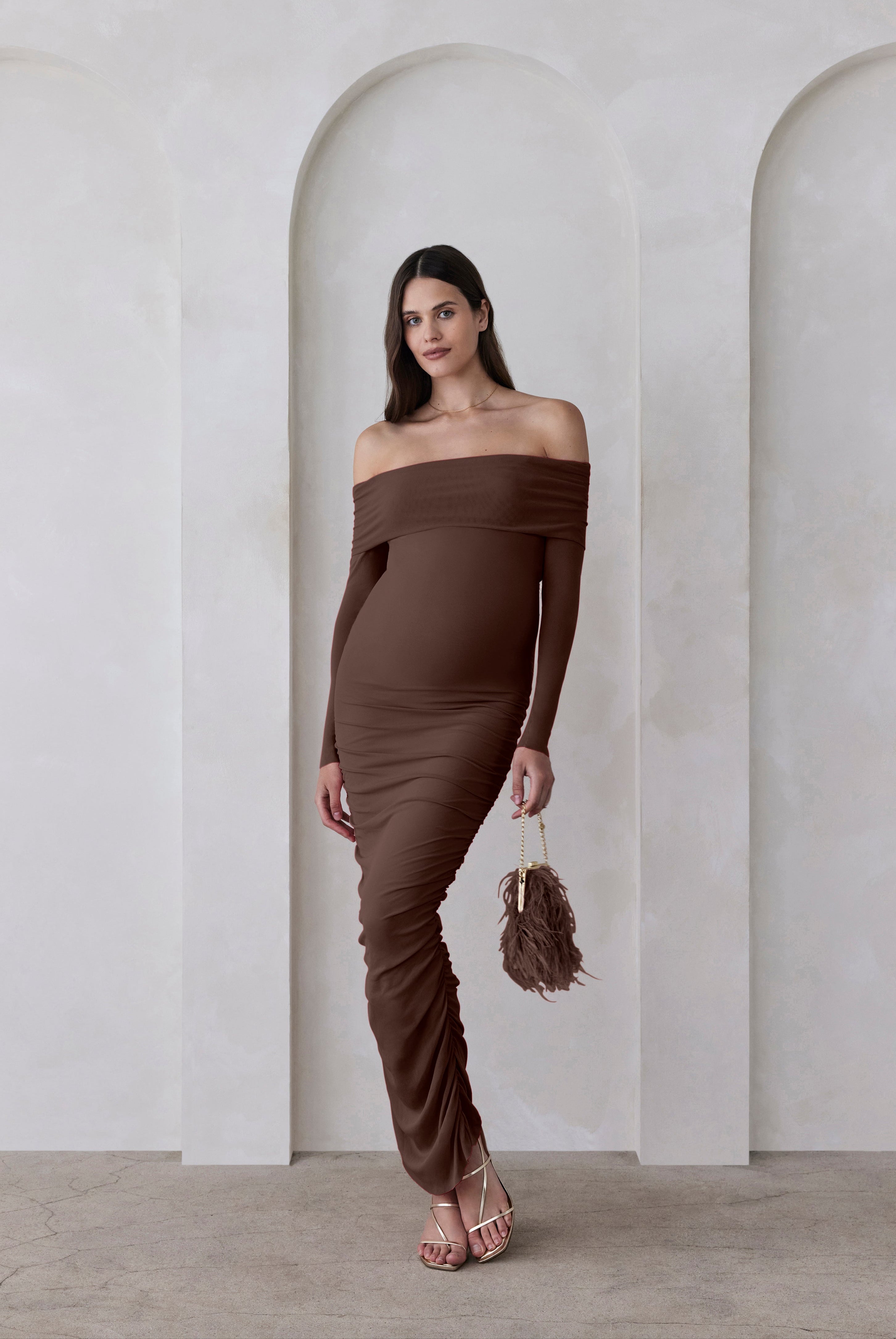 Woman wearing a brown off-shoulder dress standing in a minimalistic setting with arches.