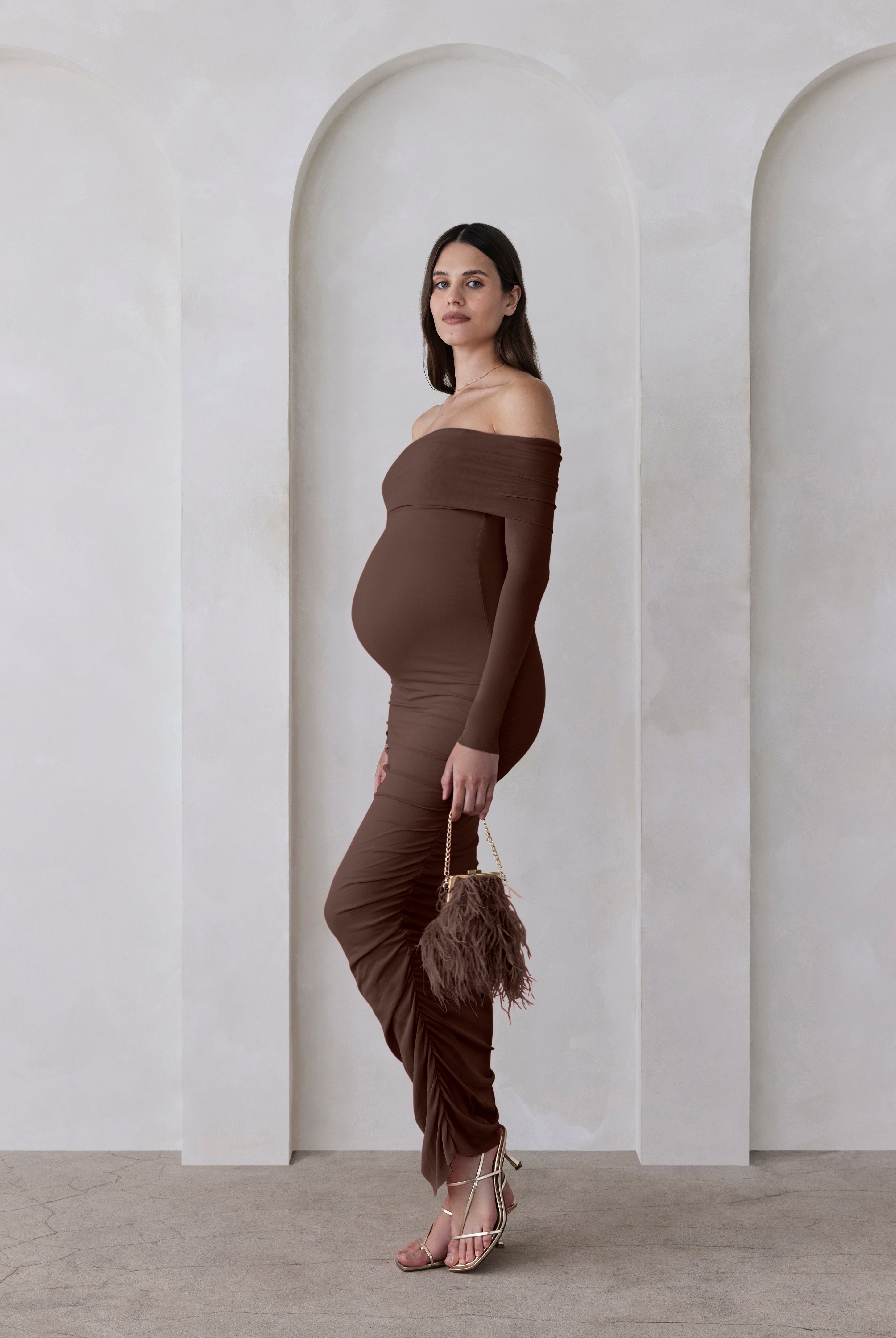 Woman in a brown off-shoulder dress standing against a white wall with arches.