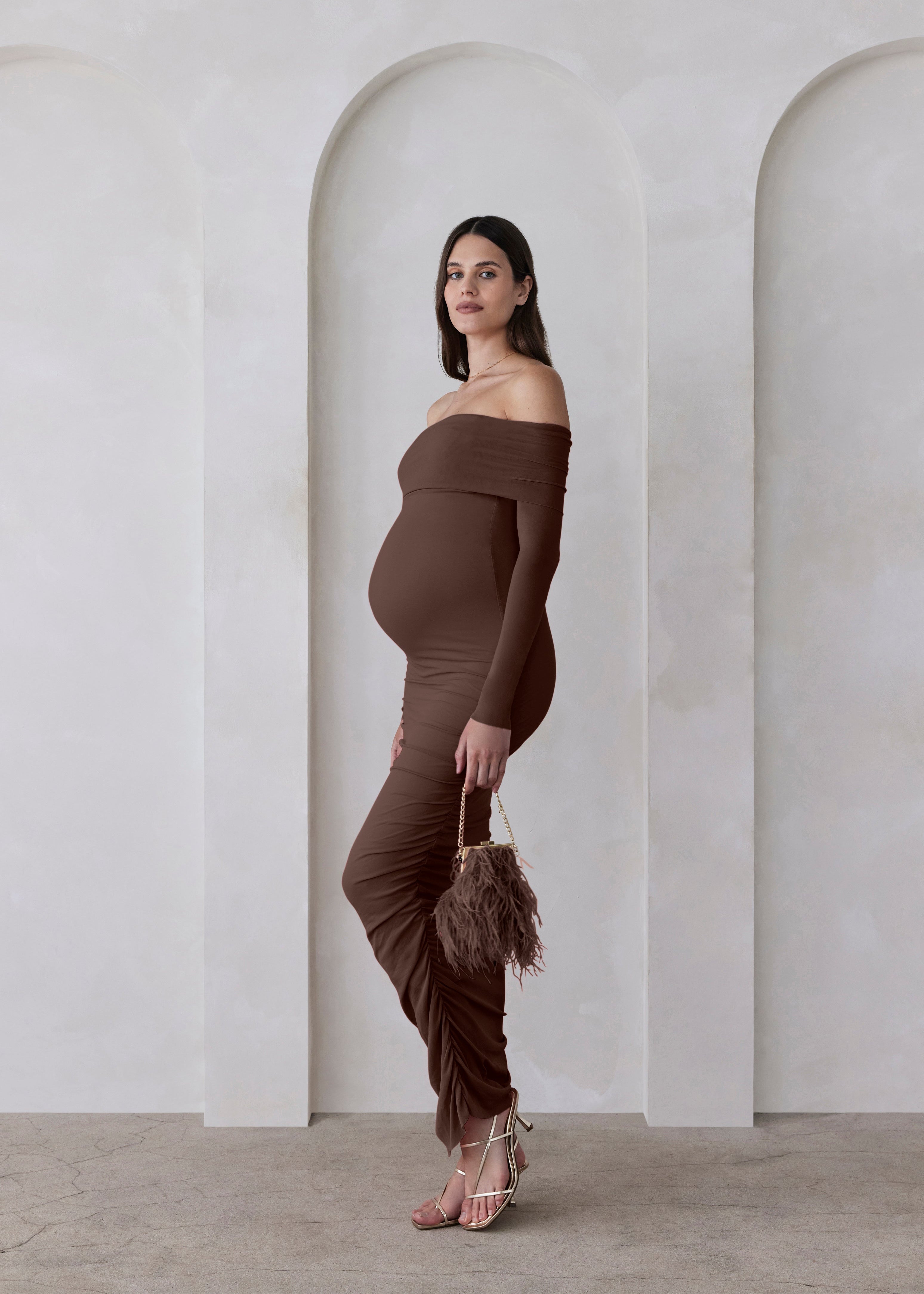 Woman in a brown off-shoulder dress standing against a white wall with arches.
