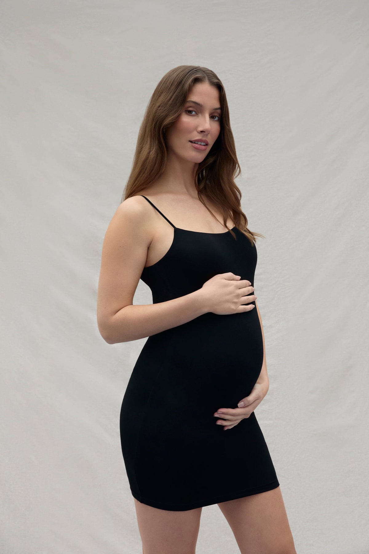 Woman holding her pregnant belly wearing a mini black dress against a plain background