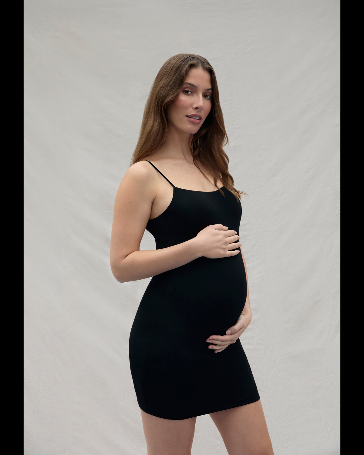 Woman holding her pregnant belly wearing a mini black dress against a plain background
