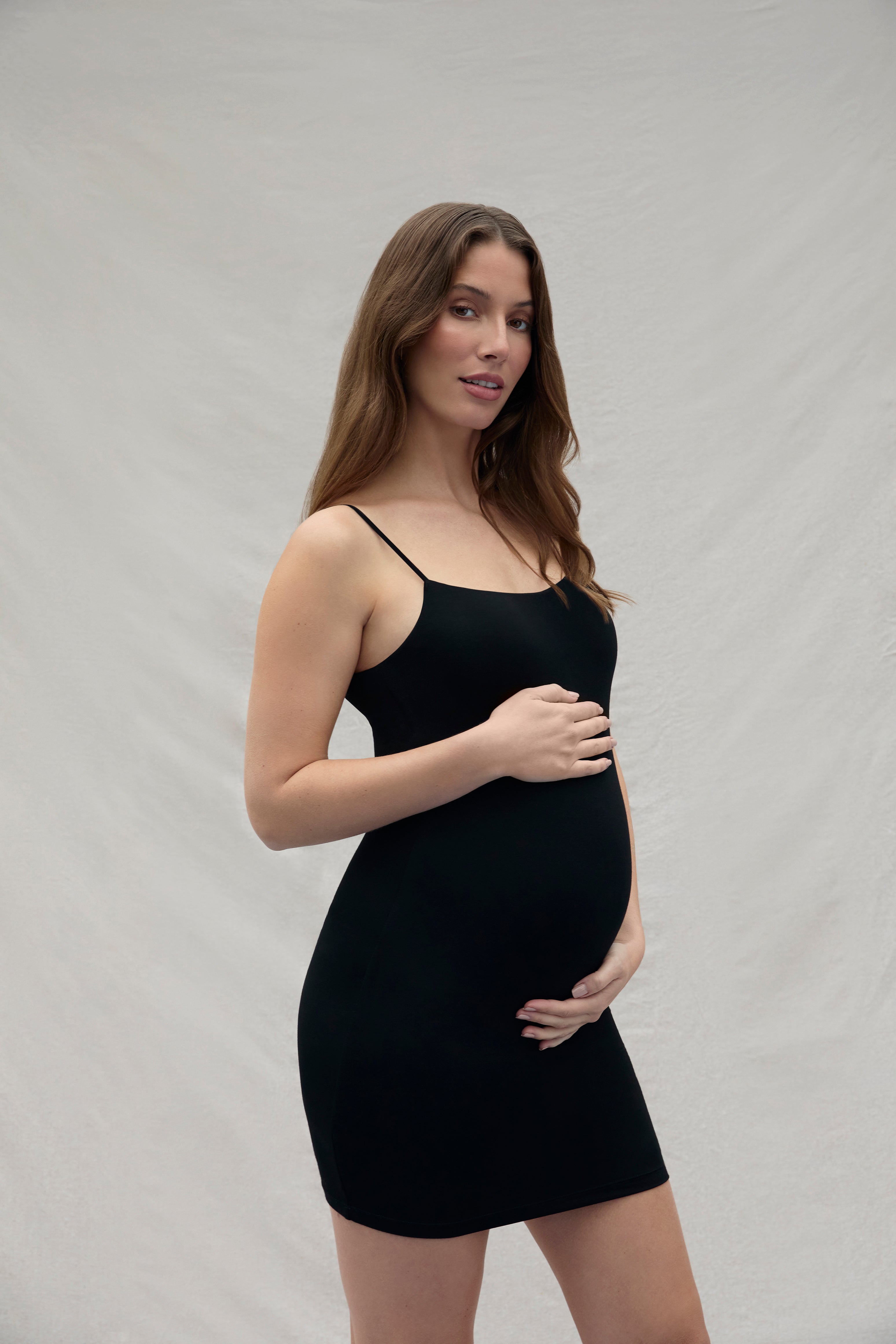 Woman holding her pregnant belly wearing a mini black dress against a plain background