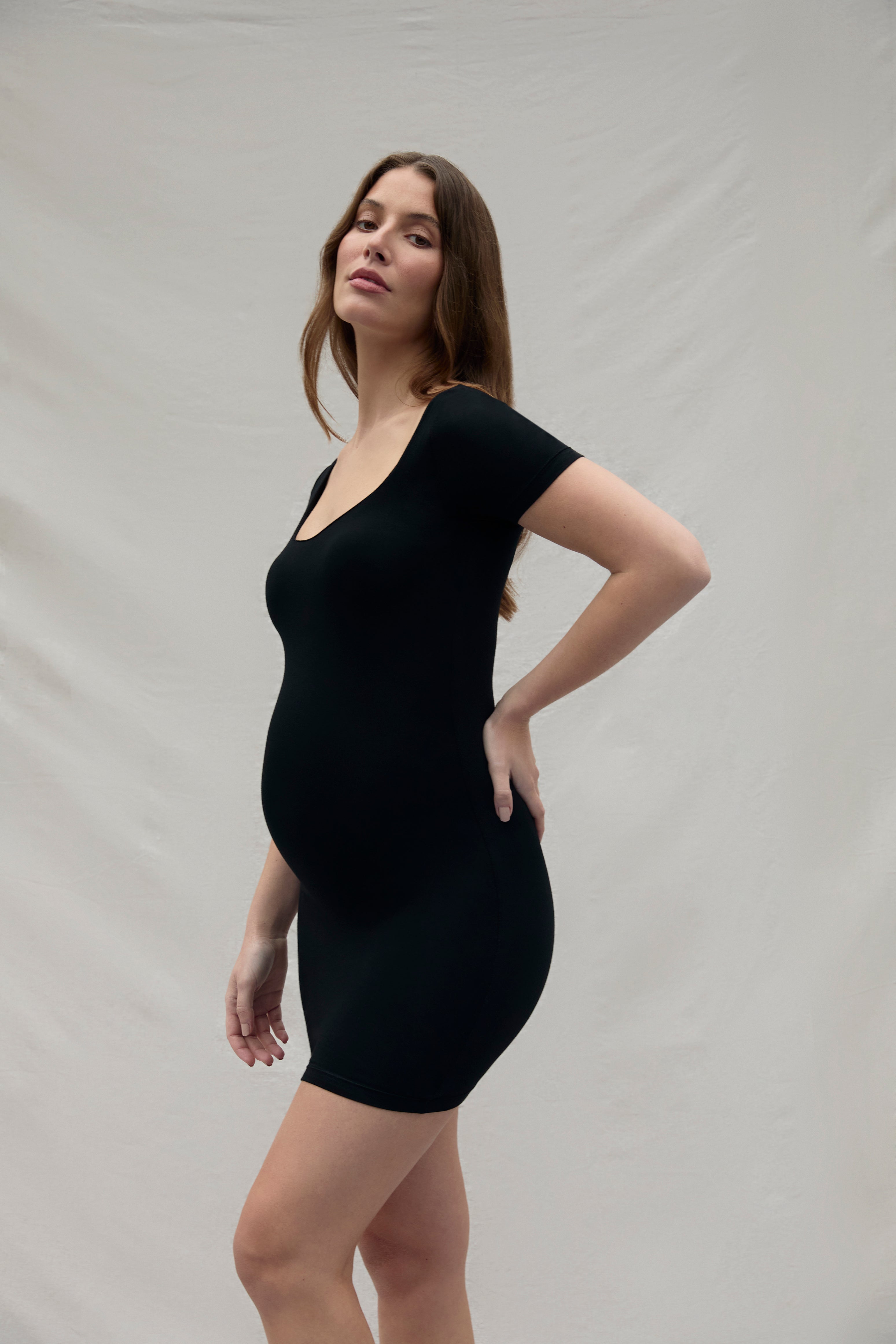 Pregnant woman wearing a black scoop neck cotton mini dress against a plain background