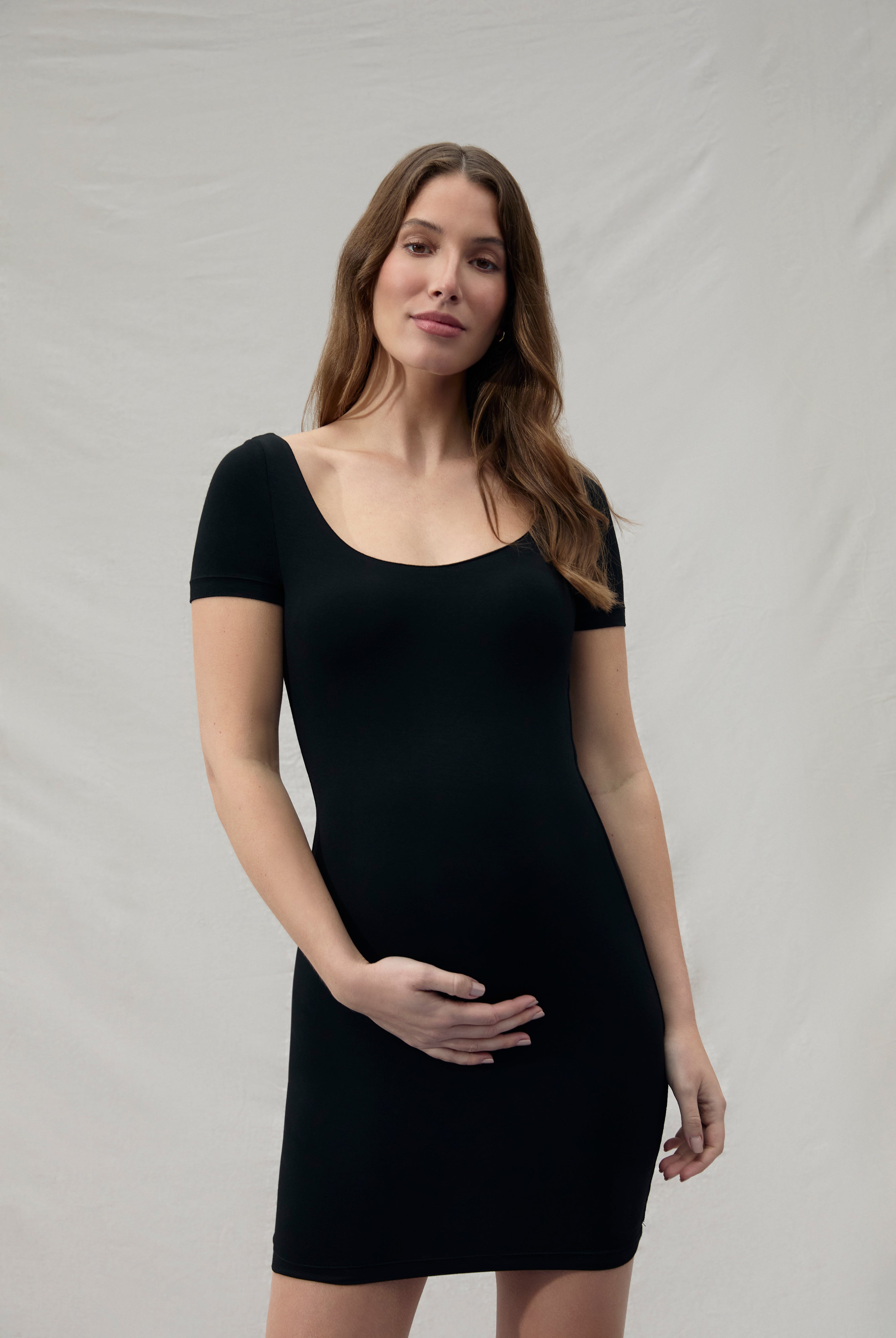 Pregnant woman wearing a mini black Bumpsuit dress against a plain background
