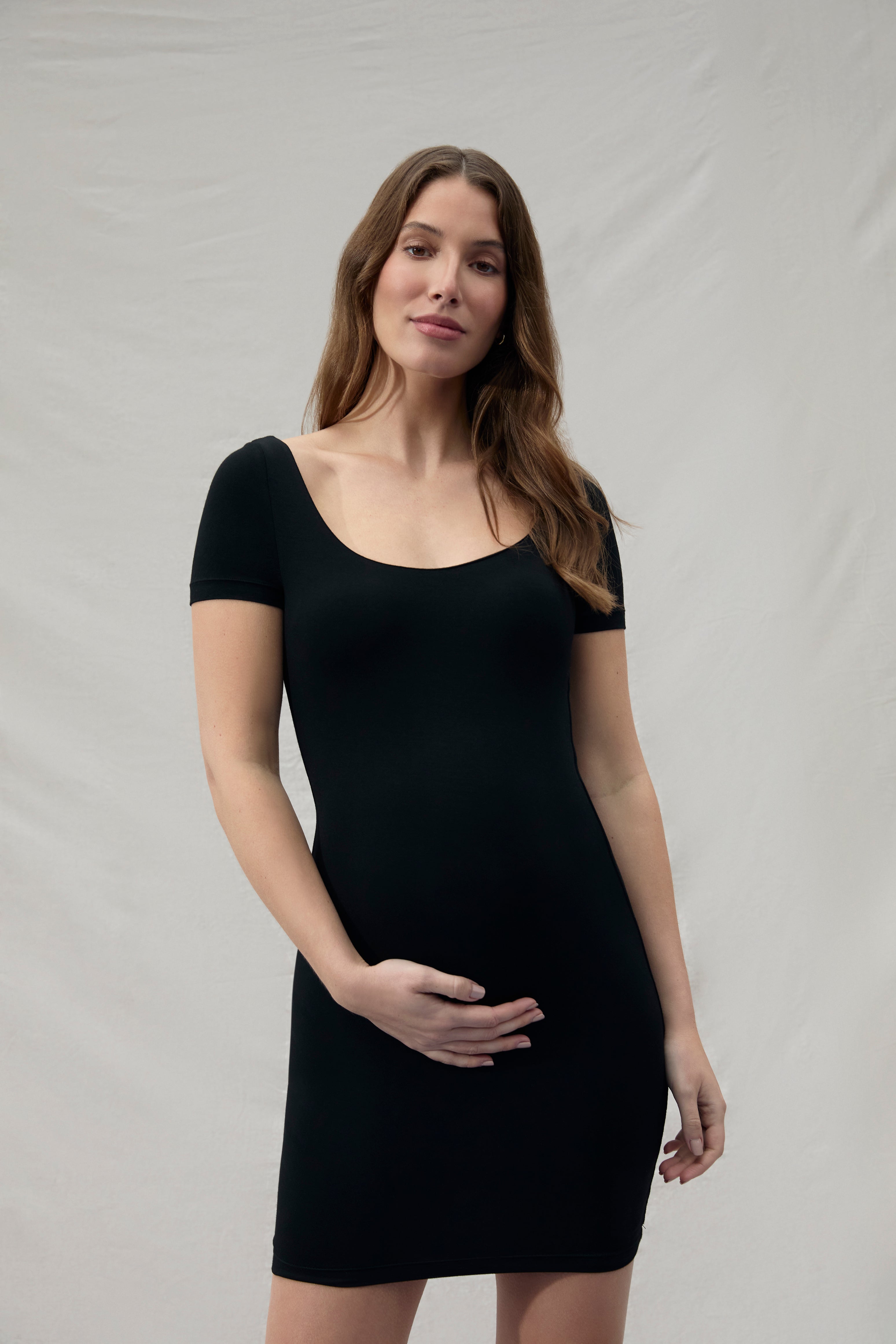 Pregnant woman wearing a mini black Bumpsuit dress against a plain background