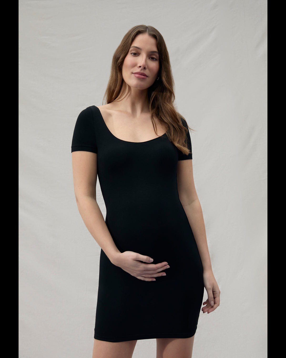 Pregnant woman wearing a mini black Bumpsuit dress against a plain background