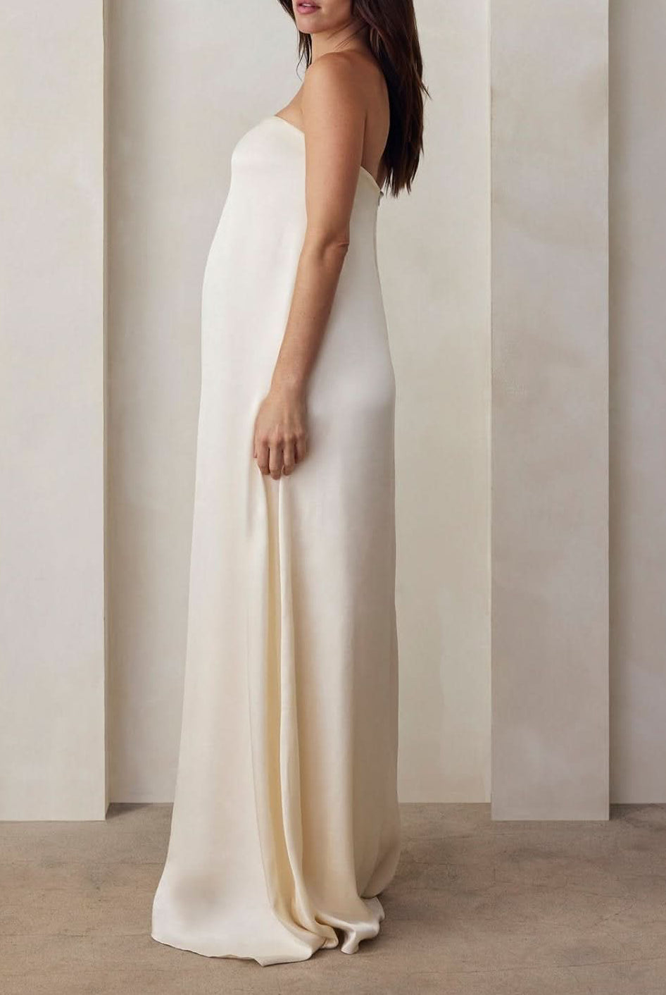 A Pregnant model wearing the Bumpsuit brushed satin gown in ivory against a beige feature wall.