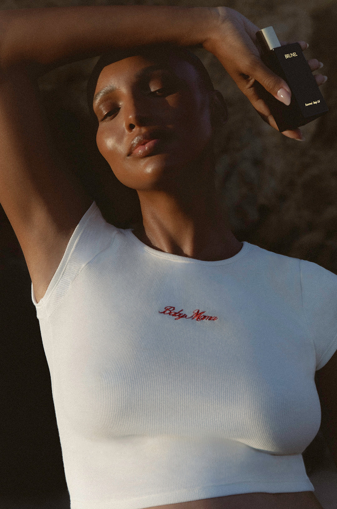 jasmine toques wearing a white bumspsuit baby mama t-shirt with red text, holding a body oil bottle against a dark background