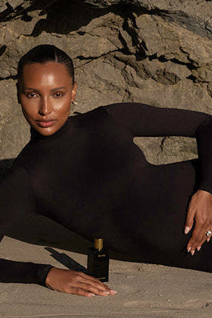 jasmine tookes in a bumpsuit black Monica dress leaning against rocks with a small bottle of body oil on the sand.