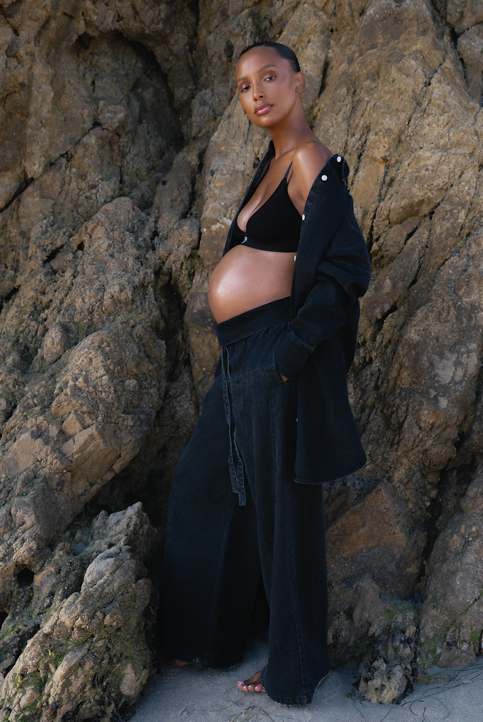 Pregnant woman in black denim Bumpsuit outfit standing against a rocky wall
