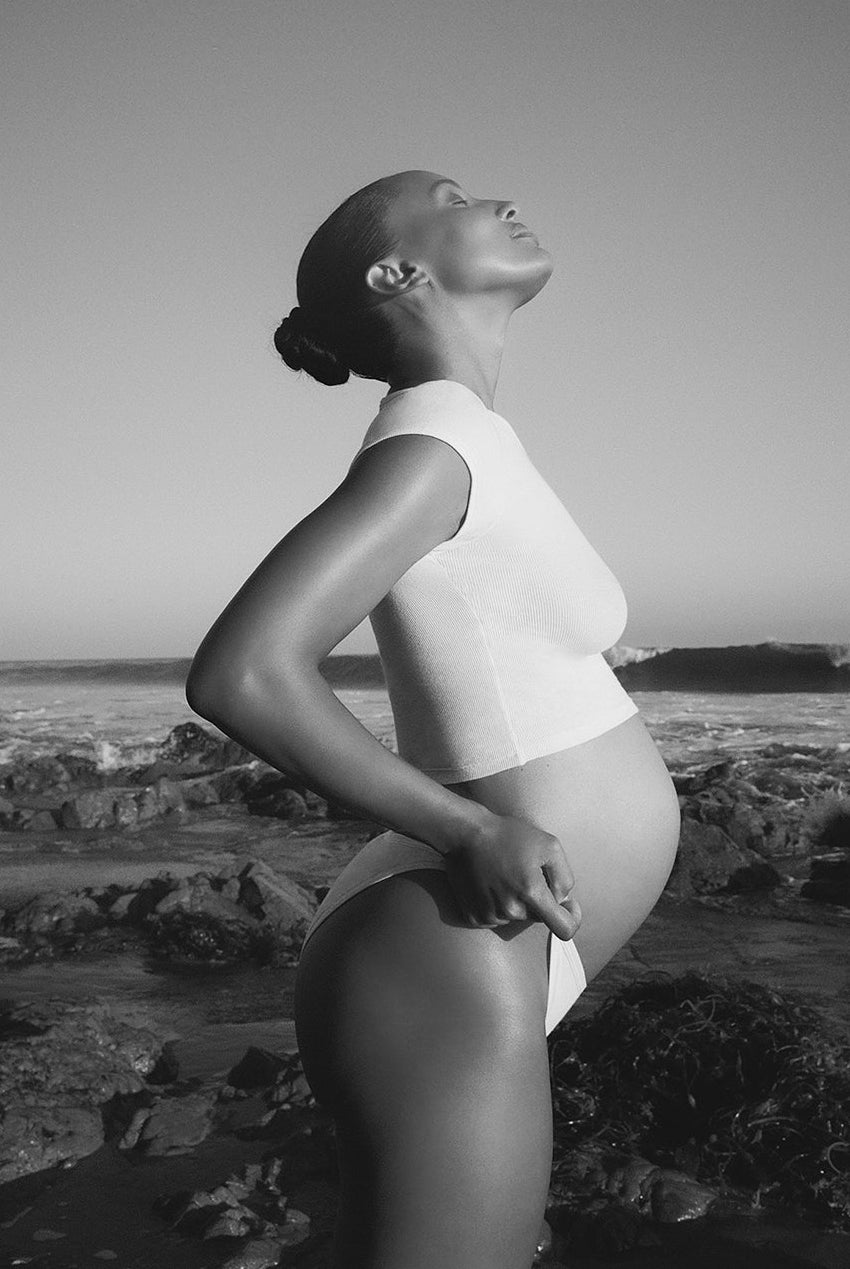Black and white photo of a pregnant woman standing on a rocky beach.