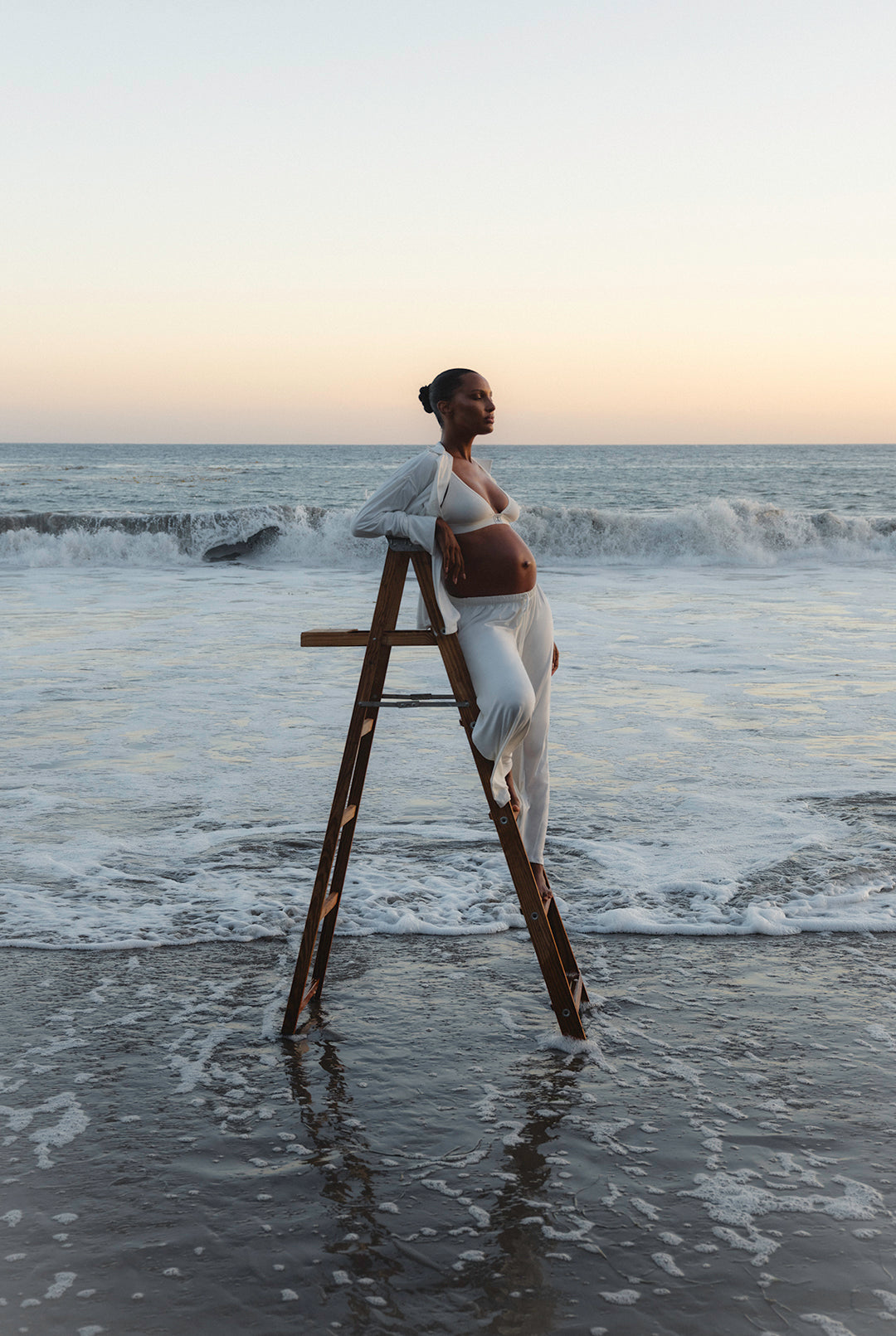 Jasmine tookes standing on a wooden ladder in the ocean with a sunset background wearing Bumpsuit Maternity Luxe Bamboo pjs in white