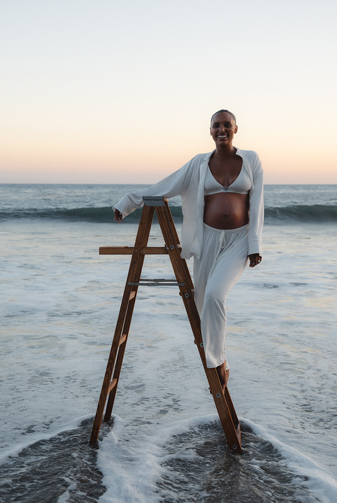 jasmine tookes standing on a ladder in the ocean wearing bumsuits maternity luxe pjs