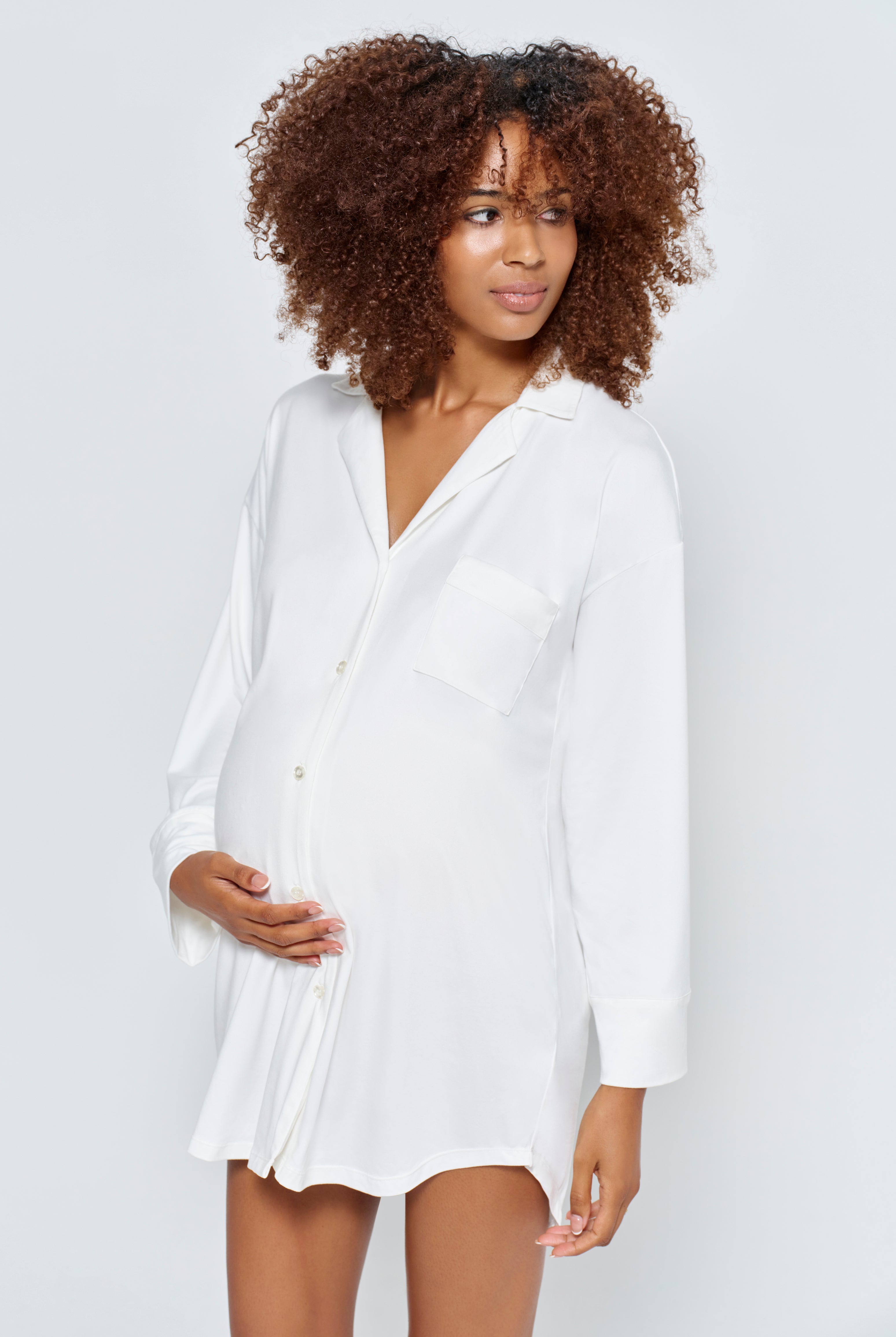 Pregnant woman wearing an ivory pyjama dress by Bumpsuit on a white background