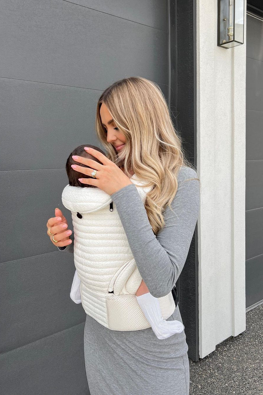 Woman wearing the Cloud Armadillo baby carrier while holding a baby outside a home