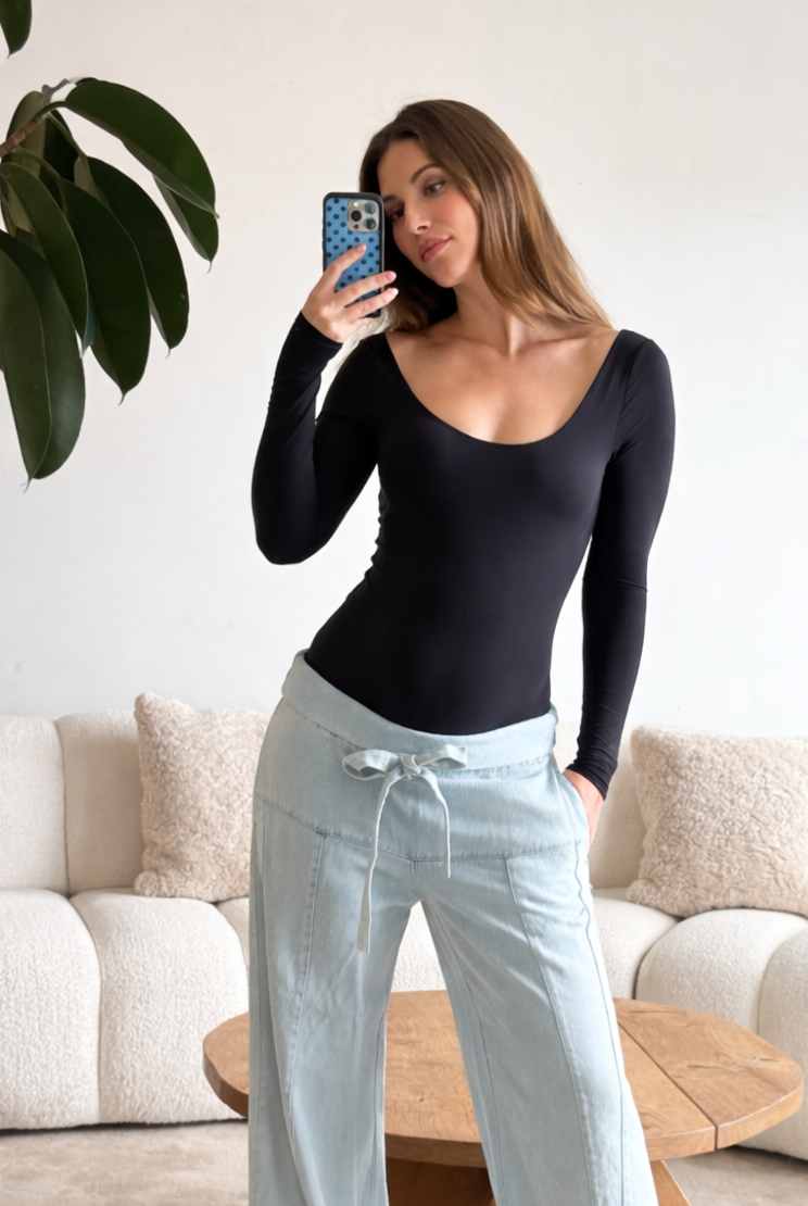 Pregnant woman taking a mirror selfie wearing bumpsuits black long-sleeve scoop neck bodysuit and light blue pants in a living room.