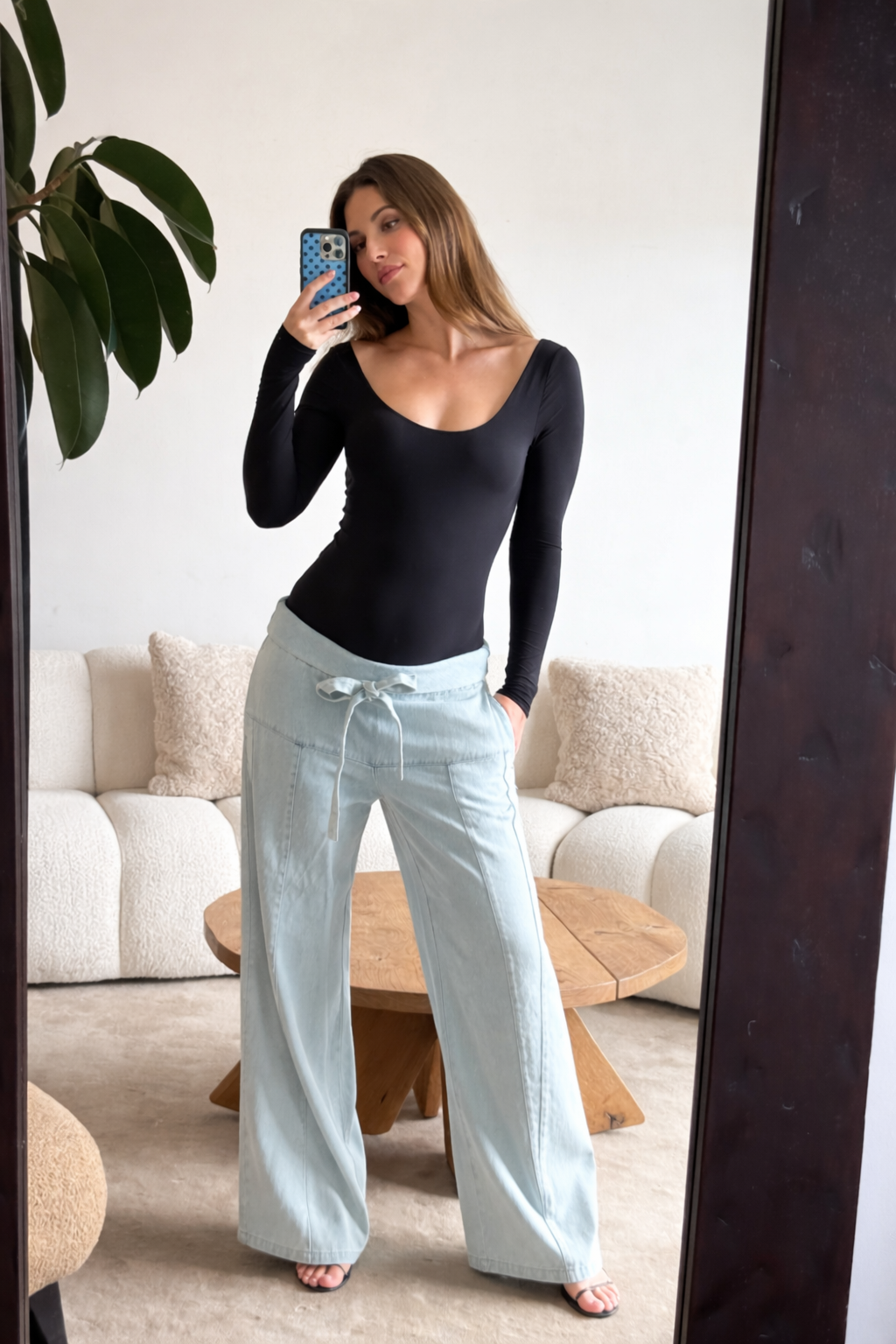 Pregnant woman taking a mirror selfie wearing bumpsuits black long-sleeve scoop neck bodysuit and light blue pants in a living room.