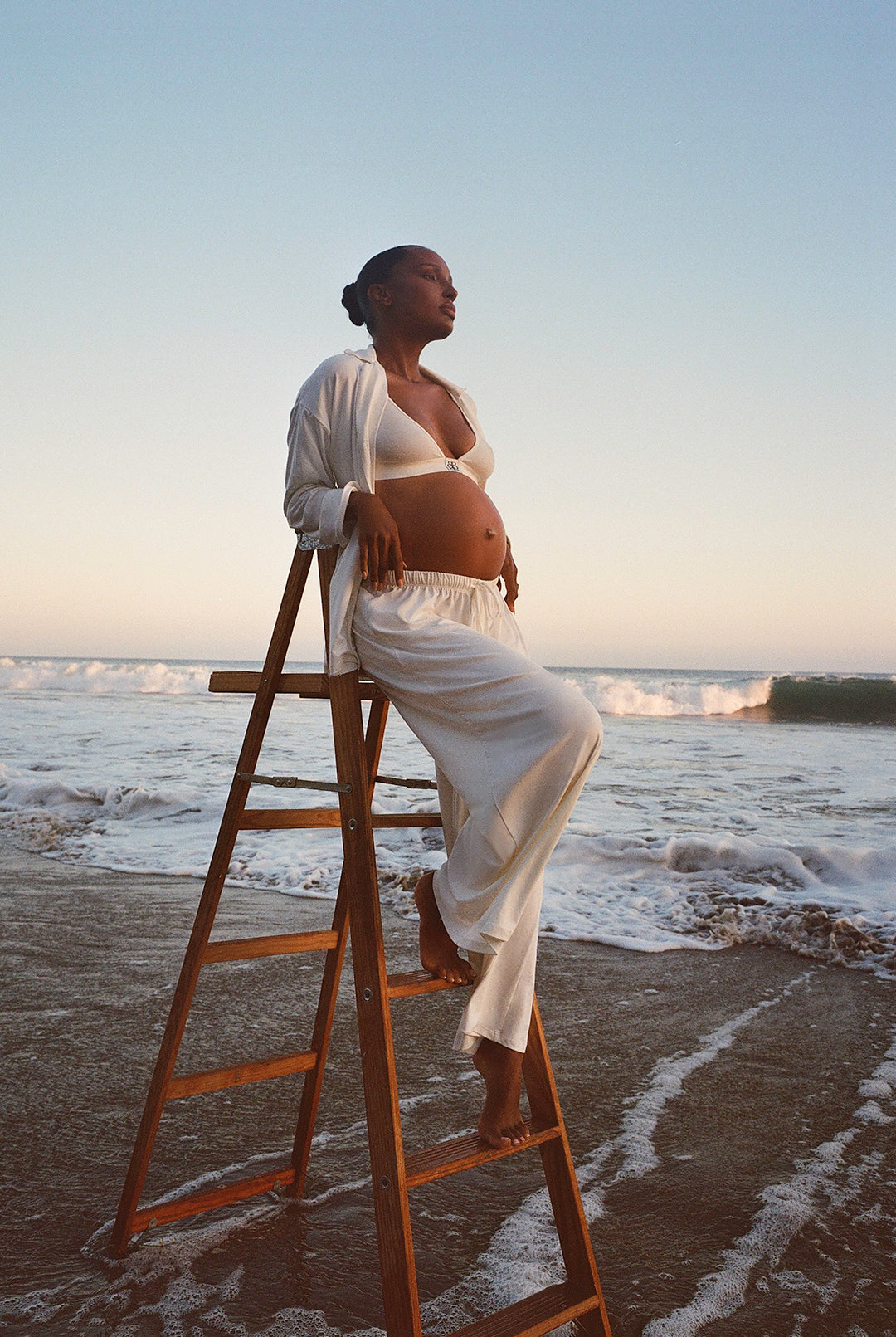 Jasmine Tookes standing on a ladder by the ocean at sunset wearing bumpsuits Luxe Bamboo Pj set