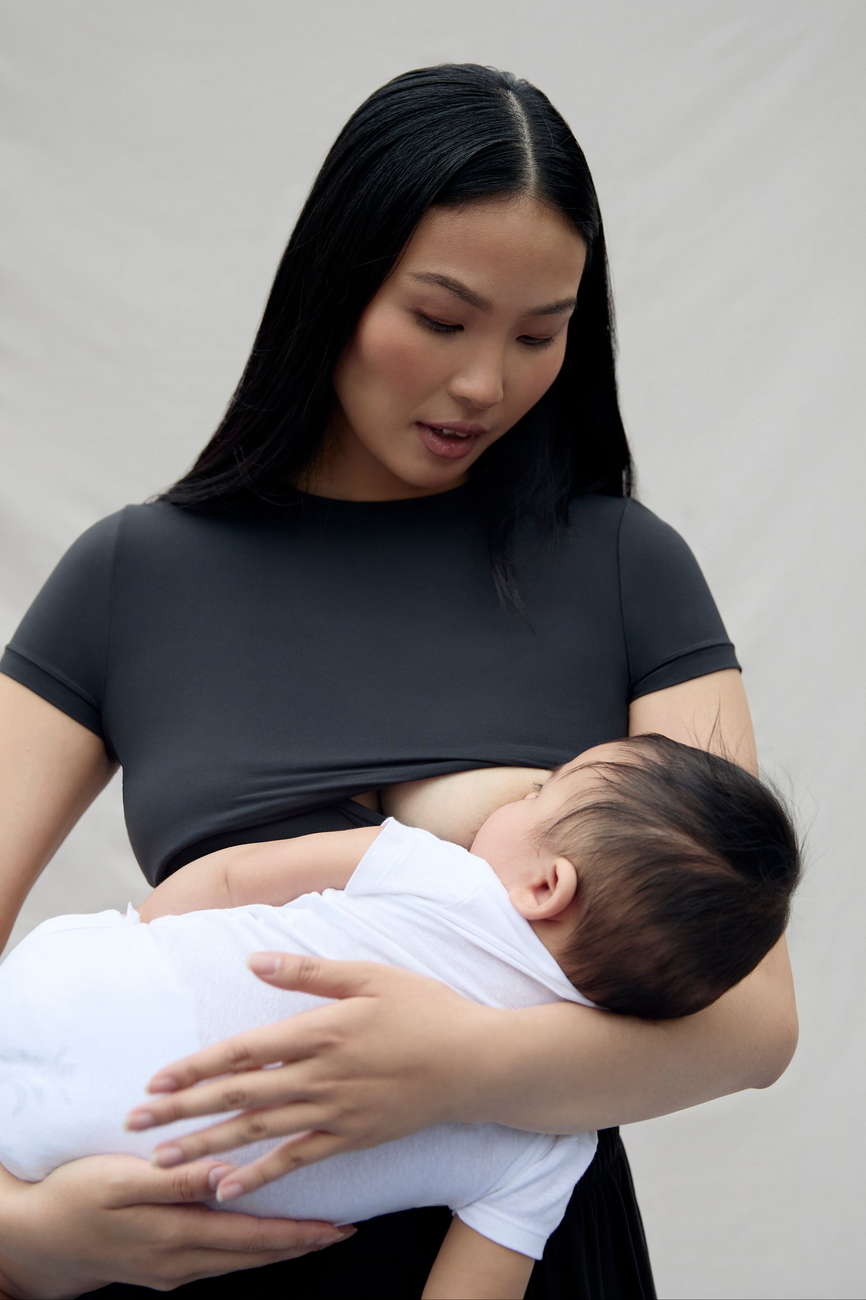 Woman feeding her baby in a black postpartum top with breast feeding access against a plain background