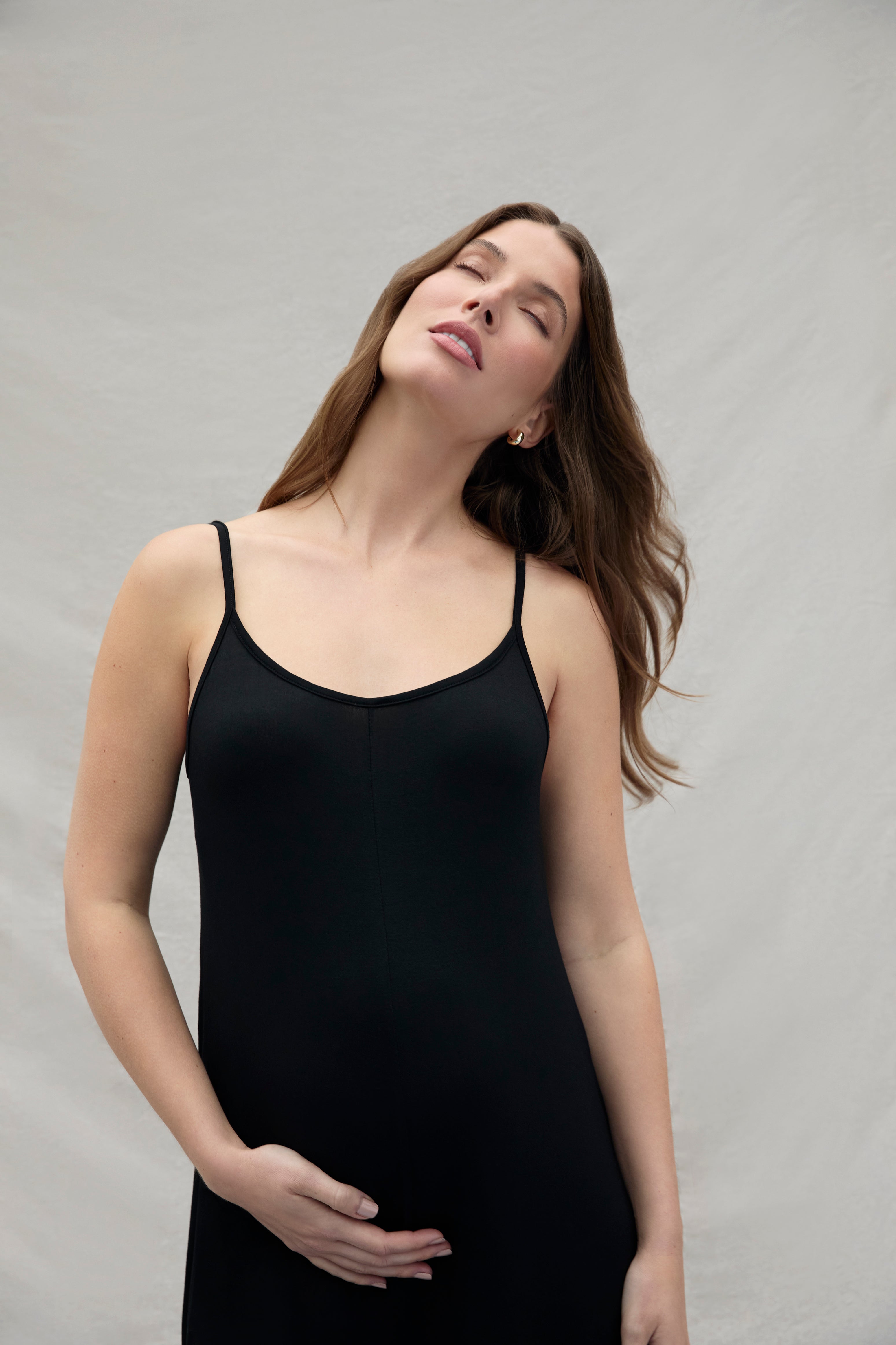Women holding under her pregnant belly wearing a cotton black jumpsuit with her eyes closed on a white background