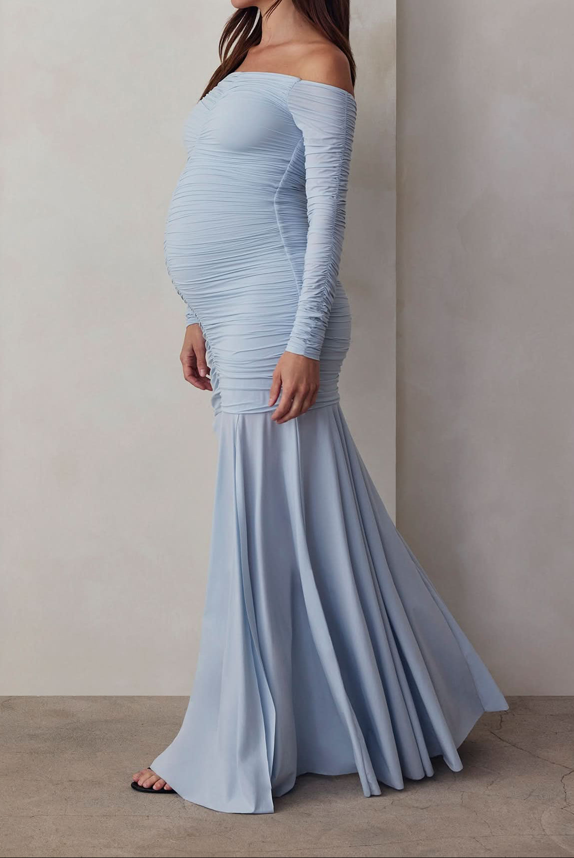 A pregnant woman wearing an off-the-shoulder, maxi-length, blue maternity dress with a flowing skirt.